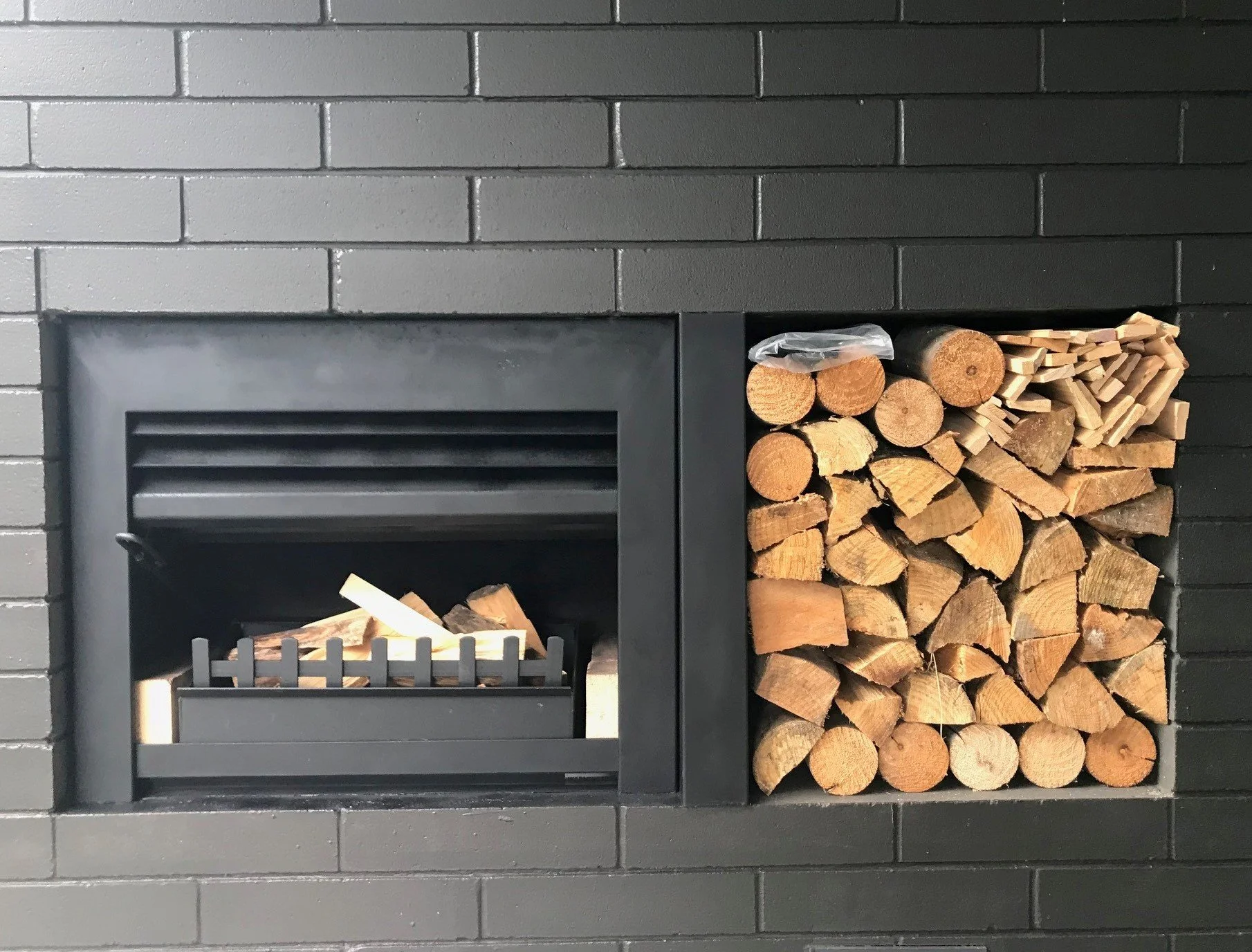 Black fireplace with a vent, stacked firewood, and small logs stored to the right.