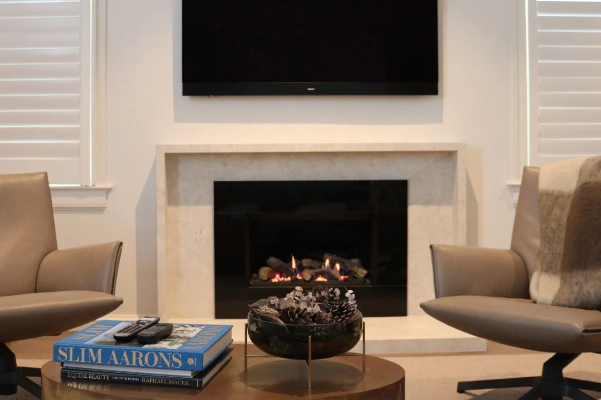 Living room with a fireplace and flat-screen TV above it, flanked by white window shutters, two beige chairs facing each other, and a coffee table with books, remotes, and decorative items.