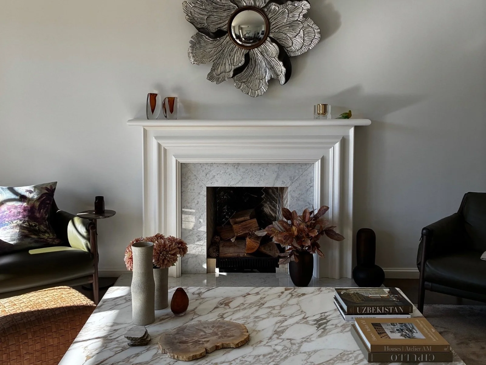 A living room with a fireplace, a marble coffee table, and decorative items. A mirror with floral metal design hangs above the fireplace. The mantel has vases and candles. Chairs with cushions and a side table are on either side of the fireplace. Books are on the coffee table.