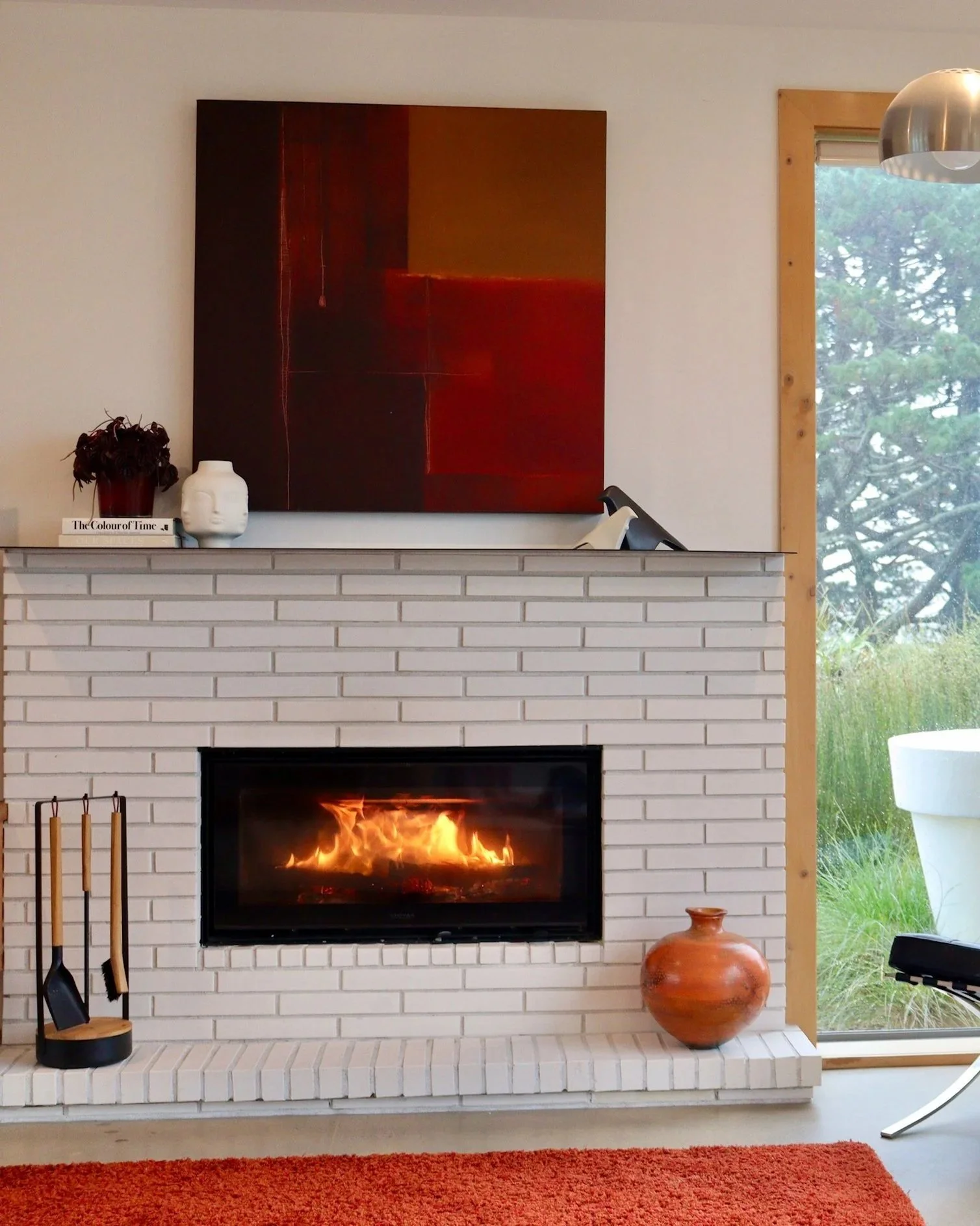 Stuv 21 wood fire in a modern living room with a built-in fireplace, wood logs stored beside it, a sectional sofa, a small wooden coffee table with magazines, a white decorative lamp, and a window seat with a cushion near a large window.