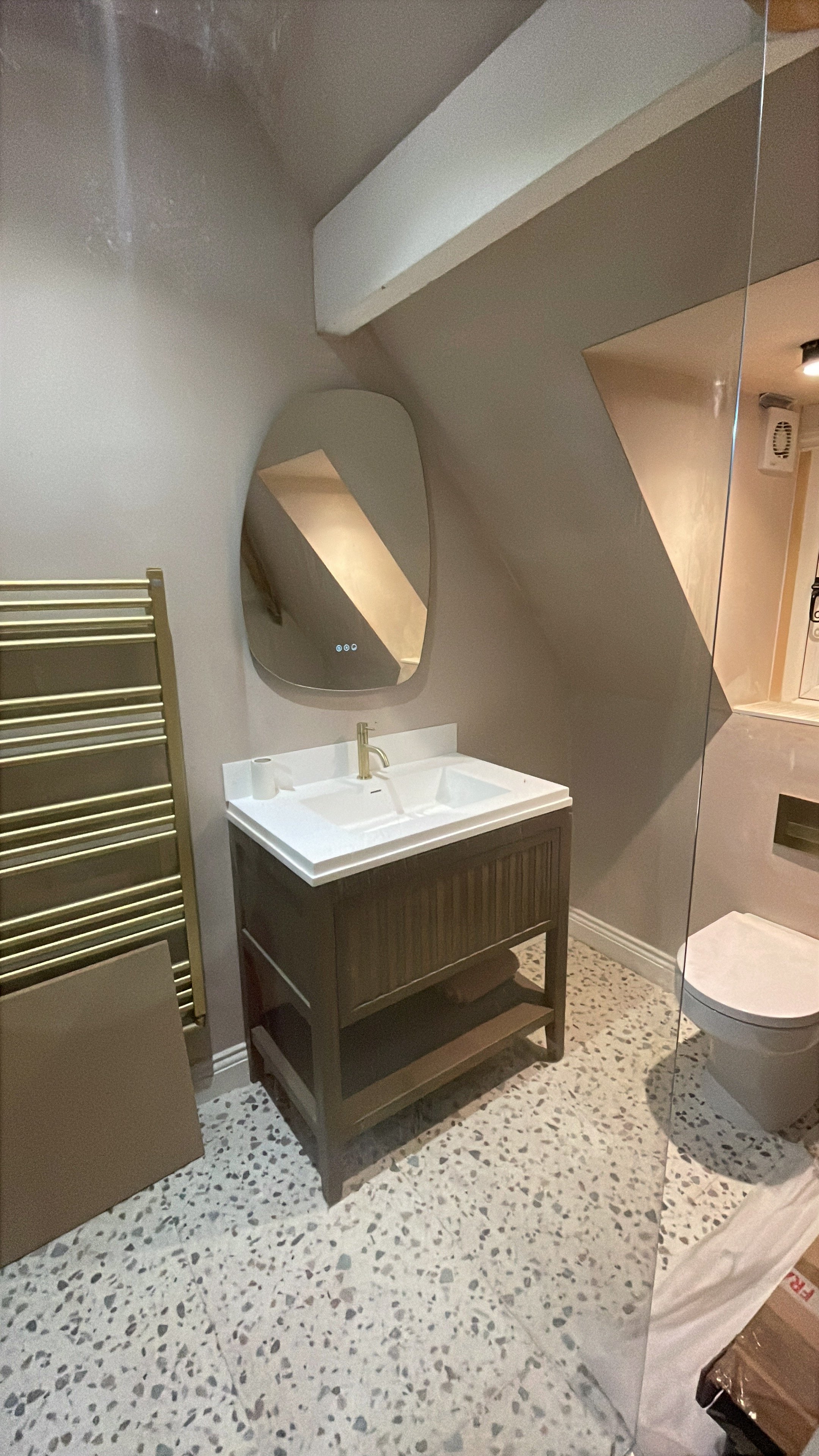 A bathroom with a small vanity sink, a mirror above it, a towel heater on the left, and a toilet on the right, with terrazzo flooring, and part of a glass shower enclosure visible.