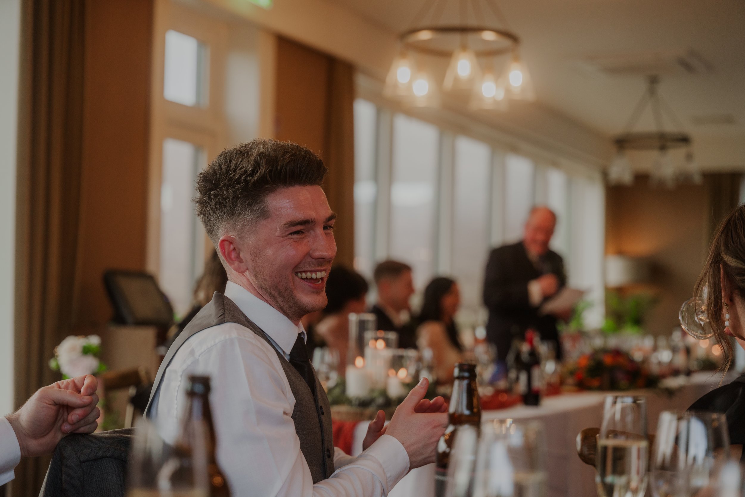 A man in a vest and tie smiling at a wedding reception, with guests and a speaker in the background.