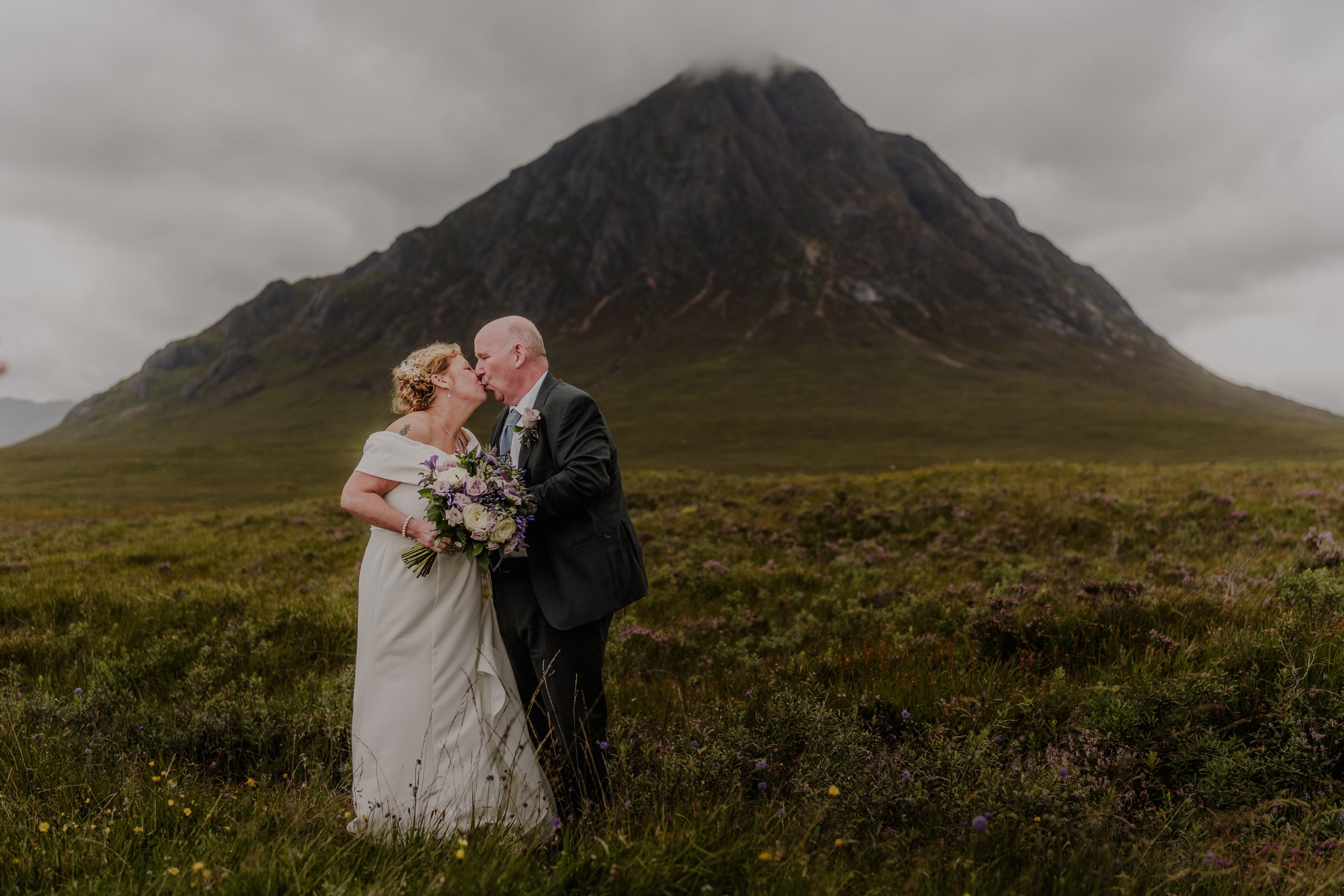 A couple in wedding attire sharing a kiss in a grassy field with a large mountain in the background, overcast sky.
