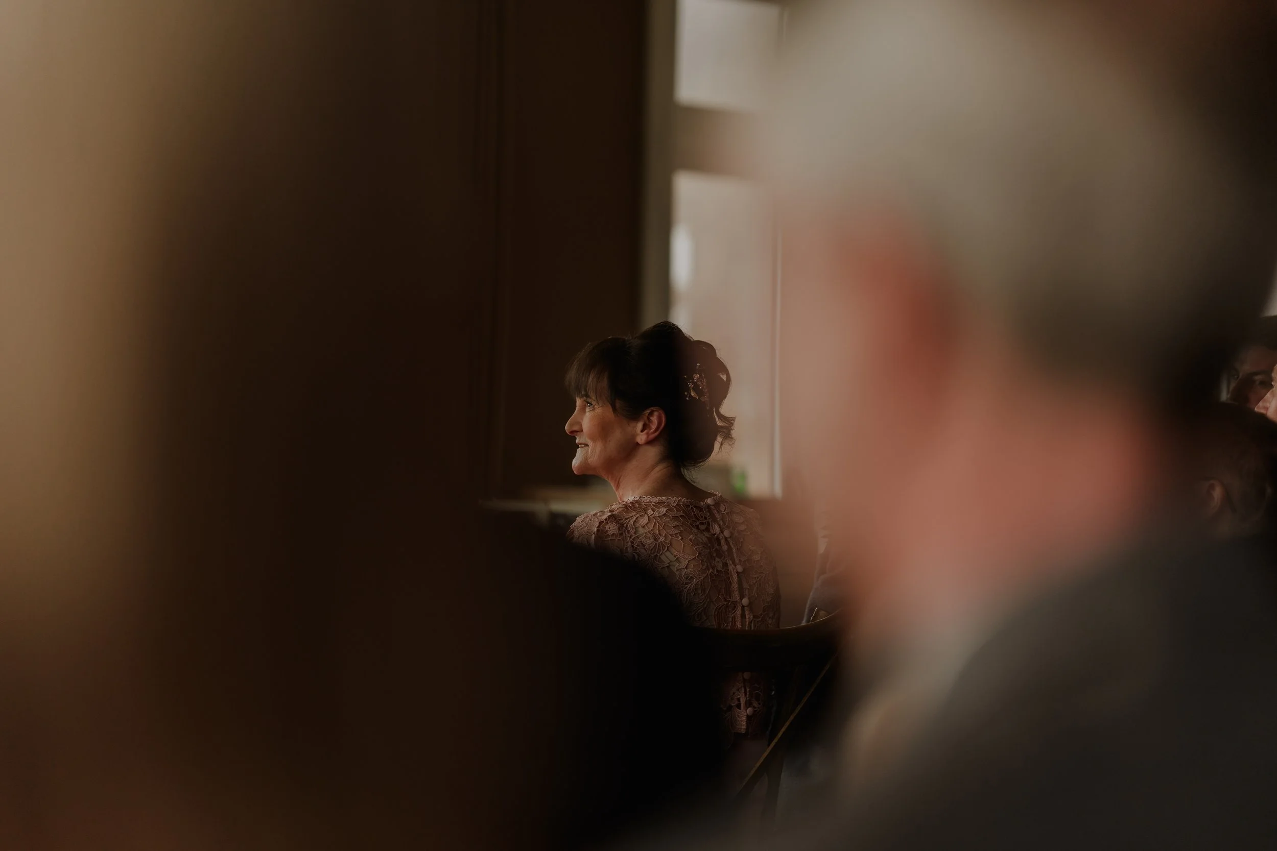 A woman with dark hair in an updo, wearing a lace dress, smiling during an indoor event, viewed through blurred foreground people.
