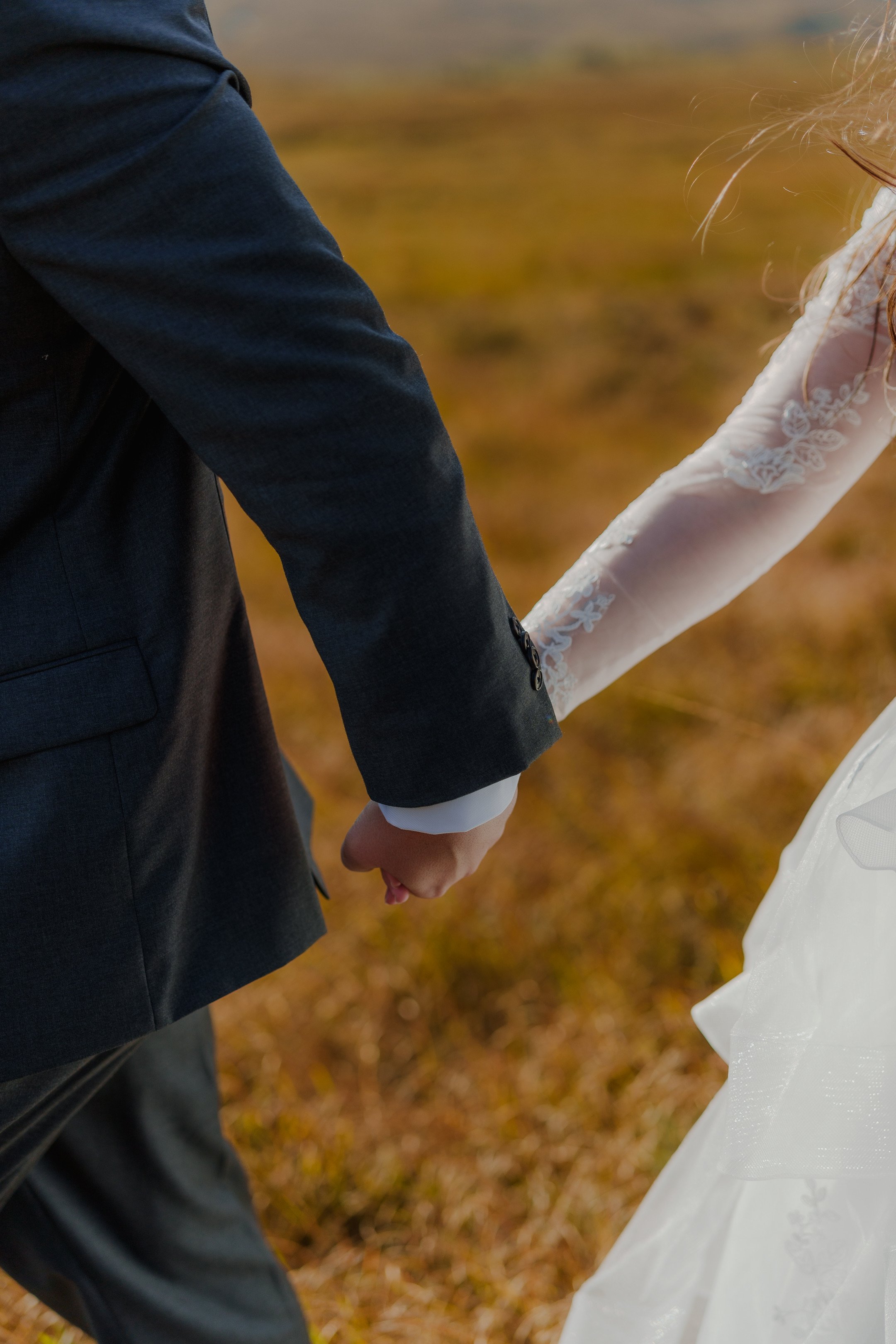 Close-up of a couple holding hands, dressed in wedding attire, standing outdoors on a fall day.