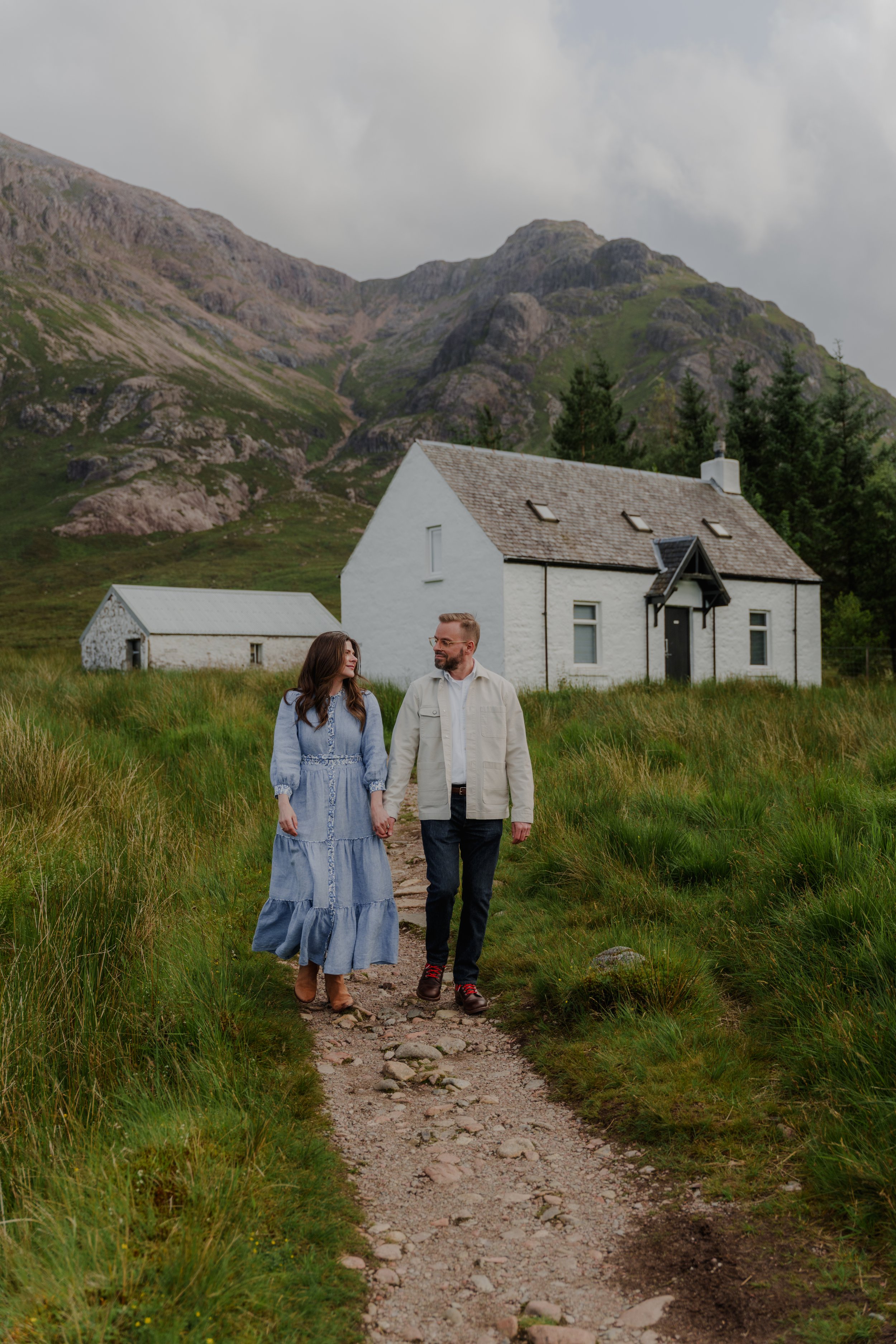 A couple holding hands walking along a dirt path in front of a white house with mountains in the background.