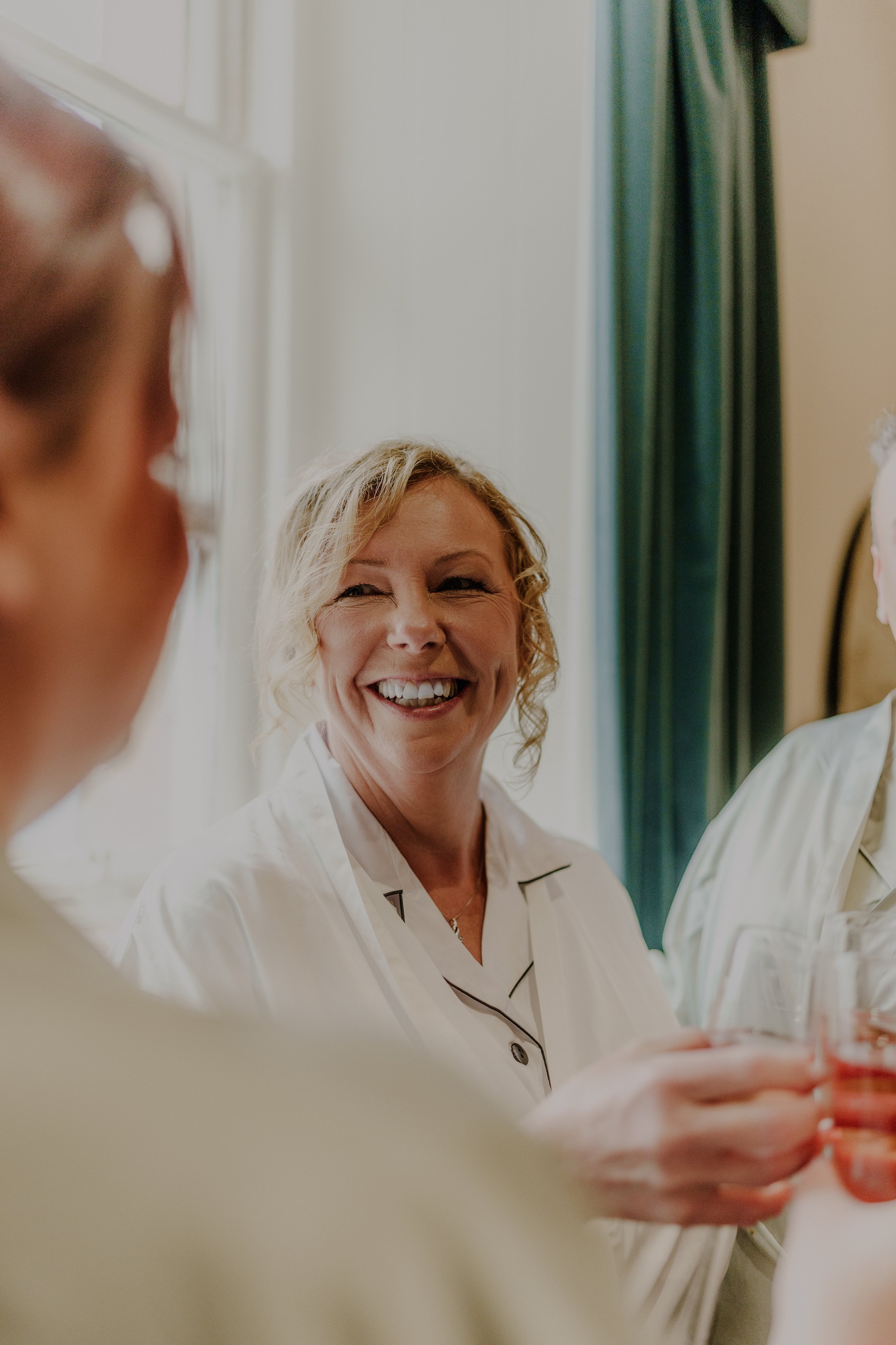 Smiling woman in pajamas holding a glass of rosé wine at a gathering with others in a bright room.