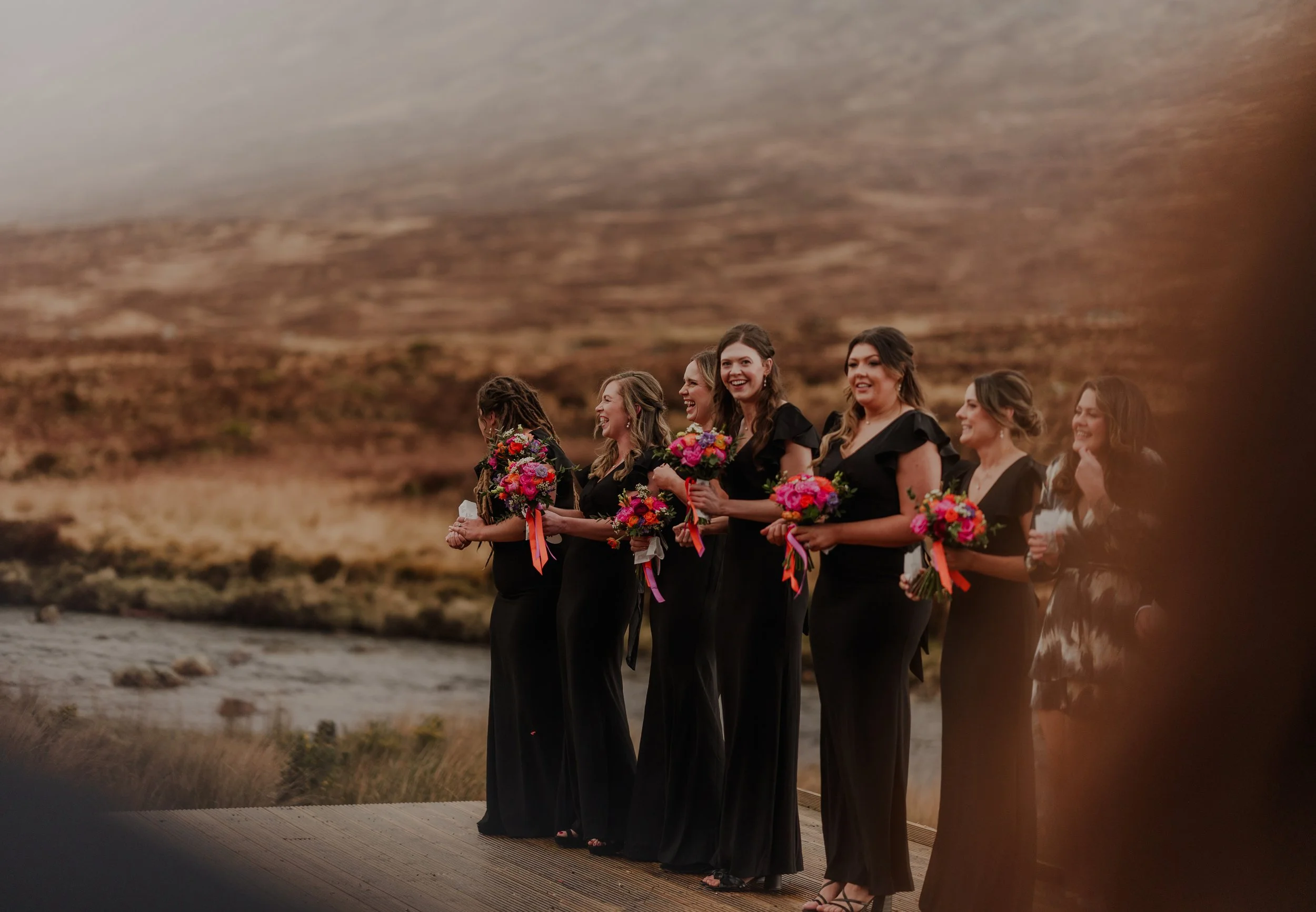 Seven women in black dresses holding colorful flower bouquets, standing outdoors with a mountain landscape in the background, smiling and laughing.