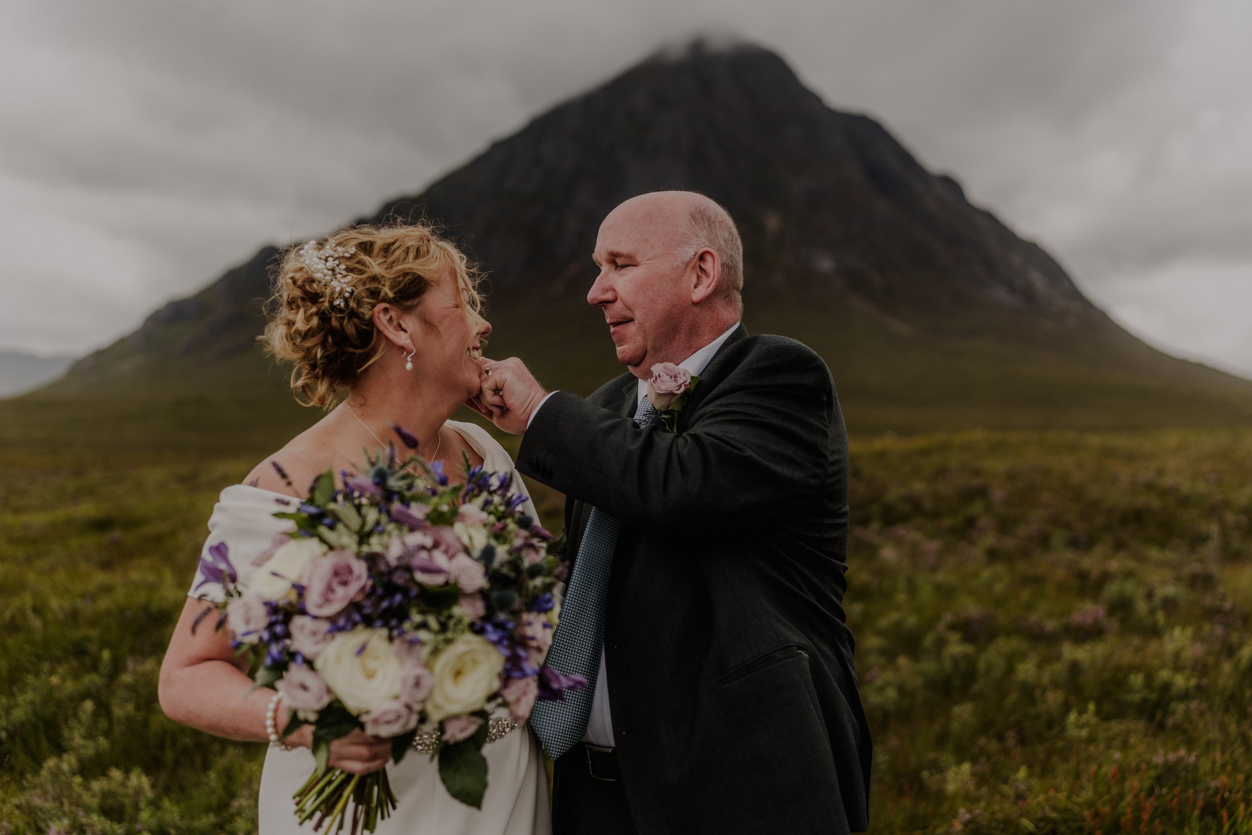 A couple dressed in wedding attire, sharing an emotional moment outdoors with a mountain in the background. The woman holds a bouquet of flowers, and the man gently touches her face.