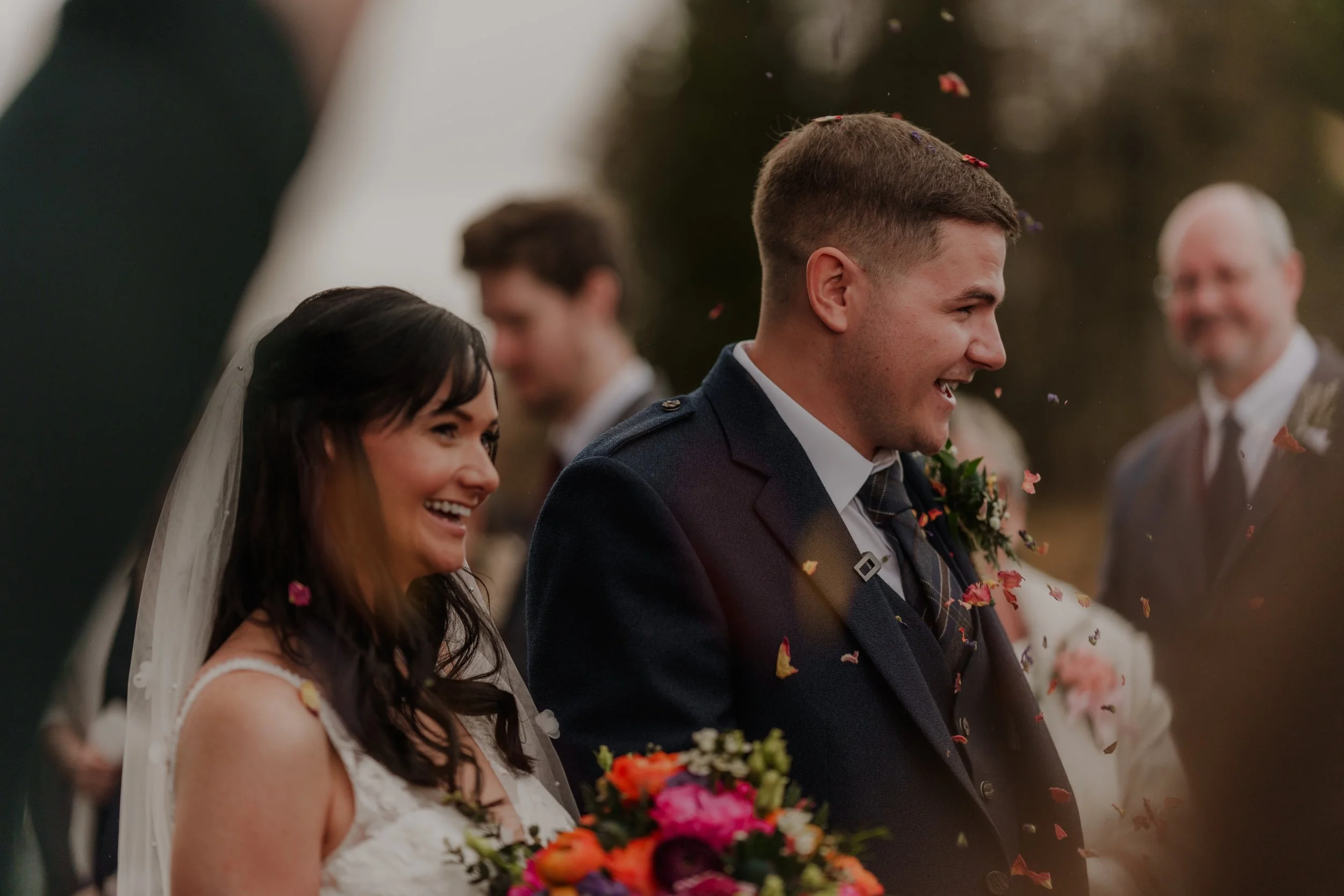 Bride and groom smiling at their wedding ceremony, surrounded by guests and falling flower petals.