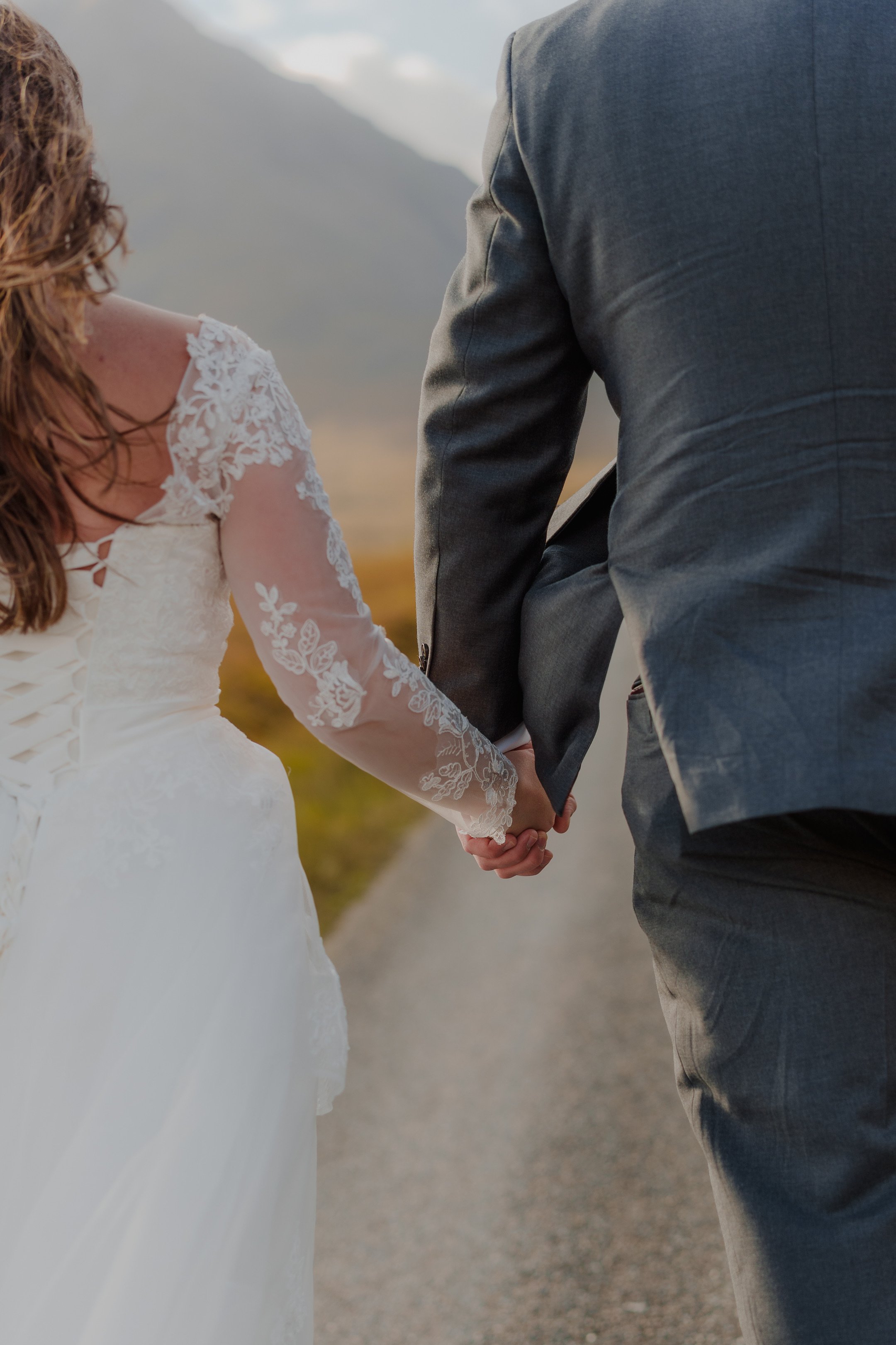 A bride and groom holding hands outdoors, walking along a rural road with mountains in the background.