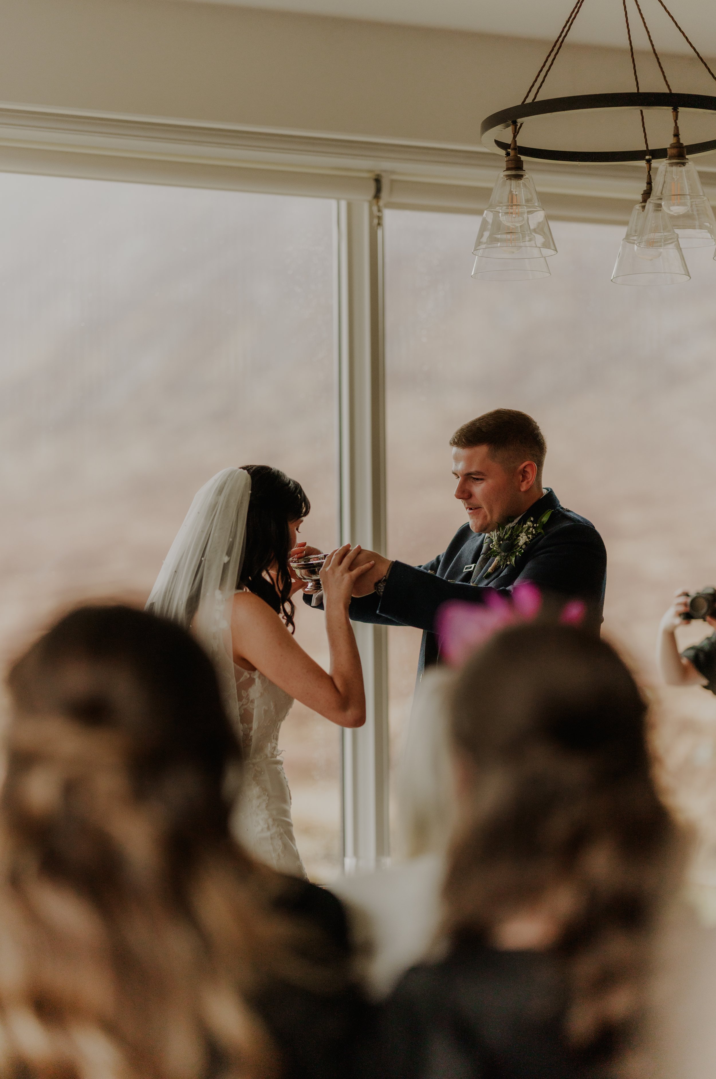 A wedding ceremony where the groom is giving the bride a drink in a small glass, in front of a large window with a view of a cloudy sky.