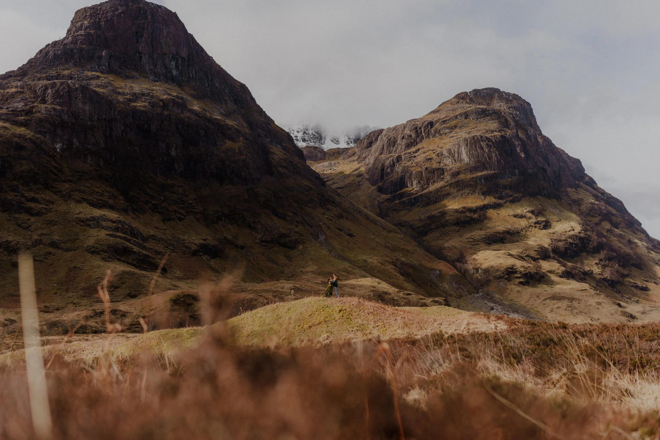 A couple standing on a grassy hill embracing each other with large mountains in the background, under a cloudy sky.