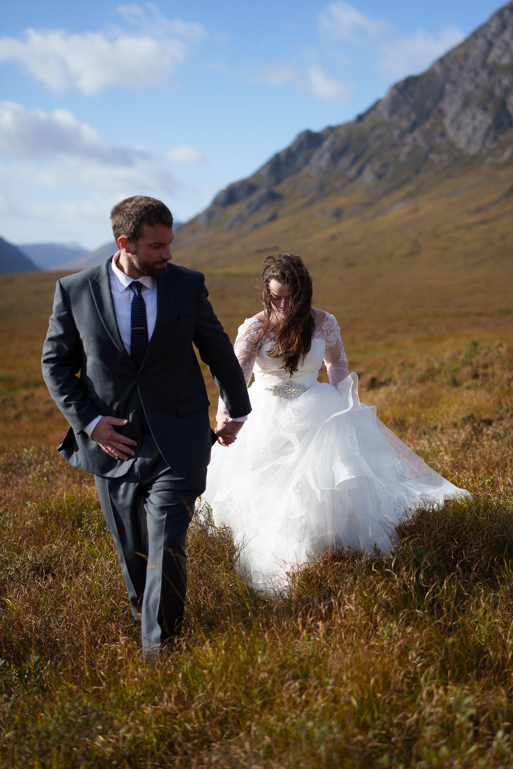 A woman in a wedding dress and a man in a suit holding hands as they walk through a grassy field with mountains in the background.