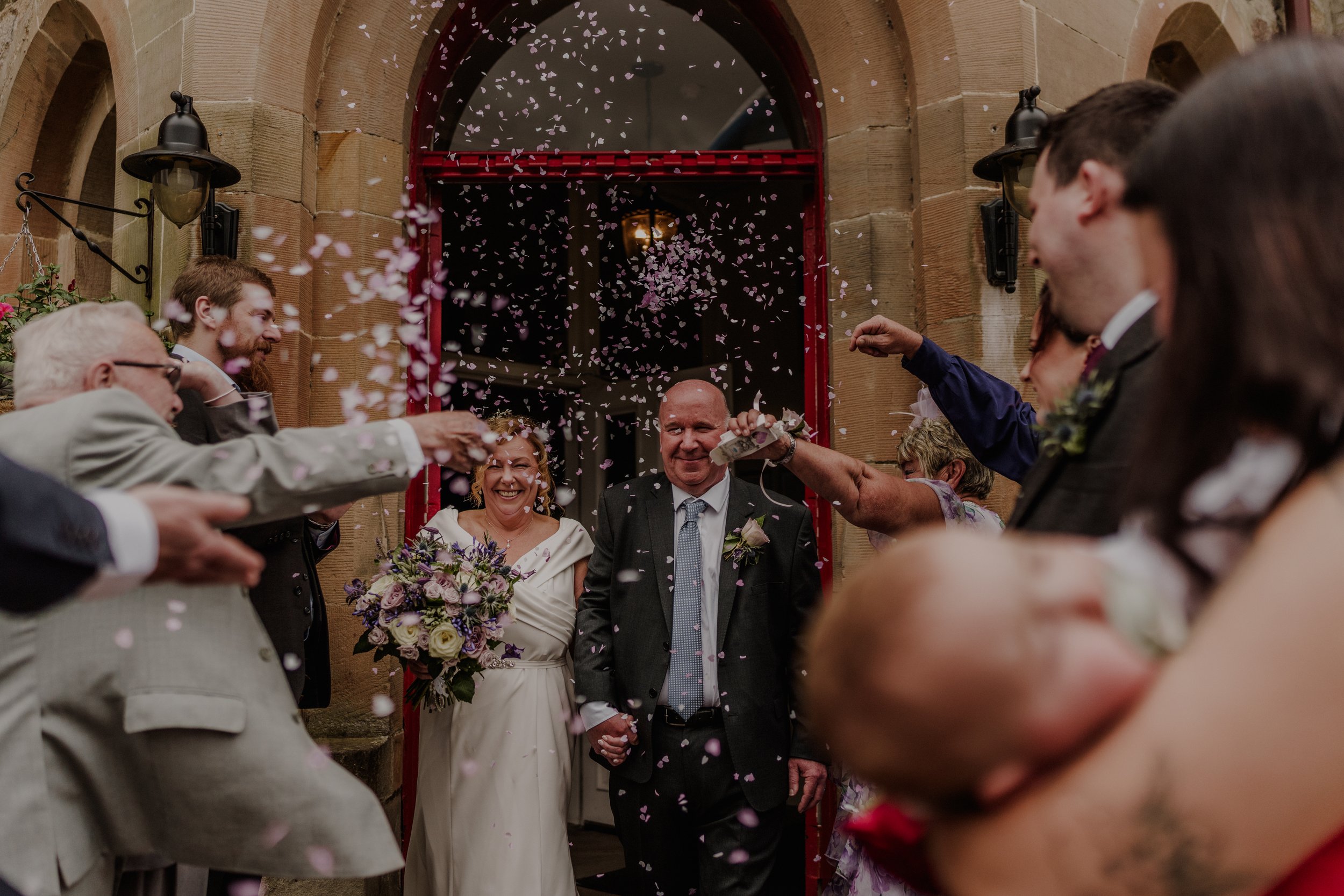 Wedding celebration with guests throwing confetti at the newly married couple in front of a building entrance.