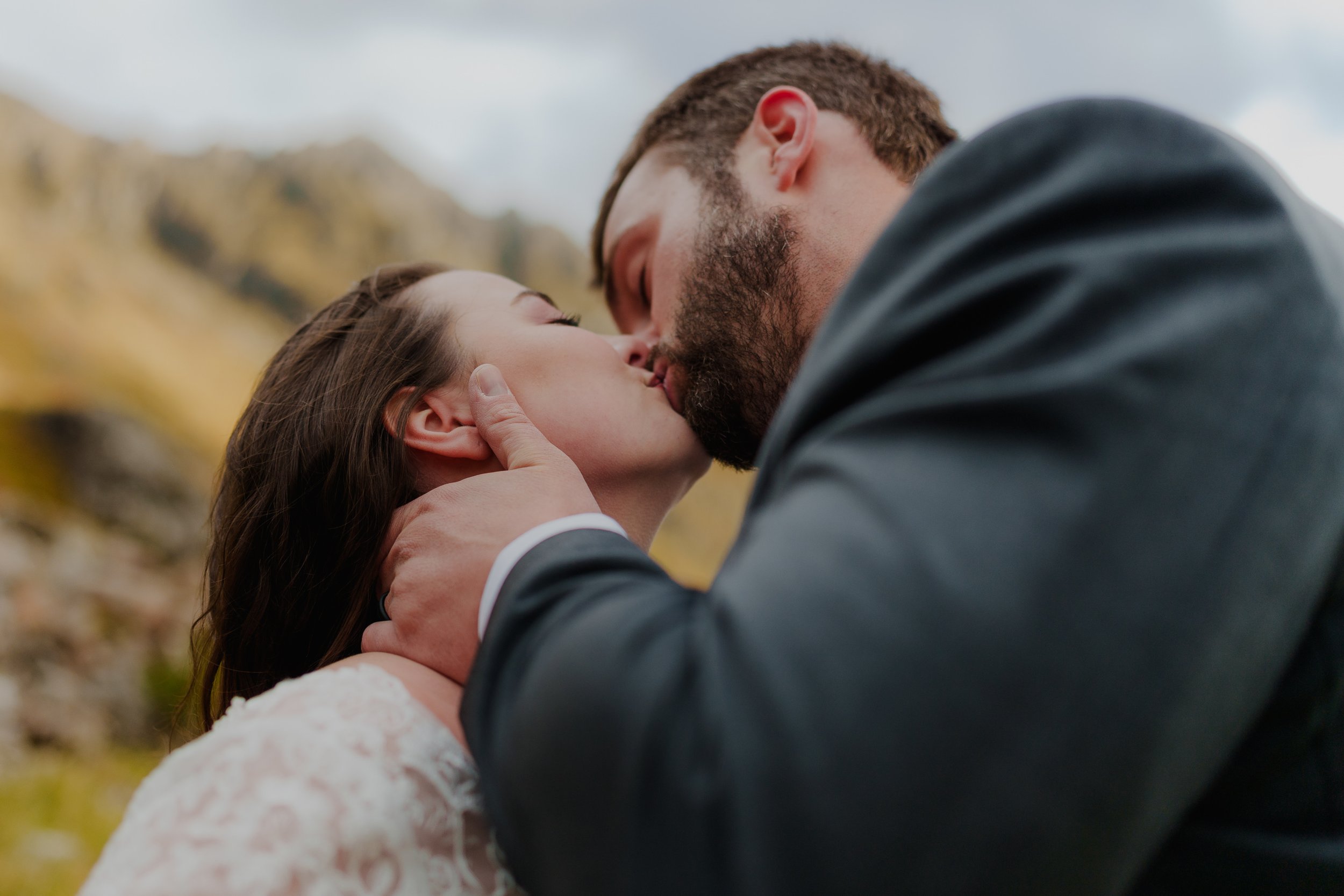 A couple sharing a kiss outdoors, with a blurred mountain background.