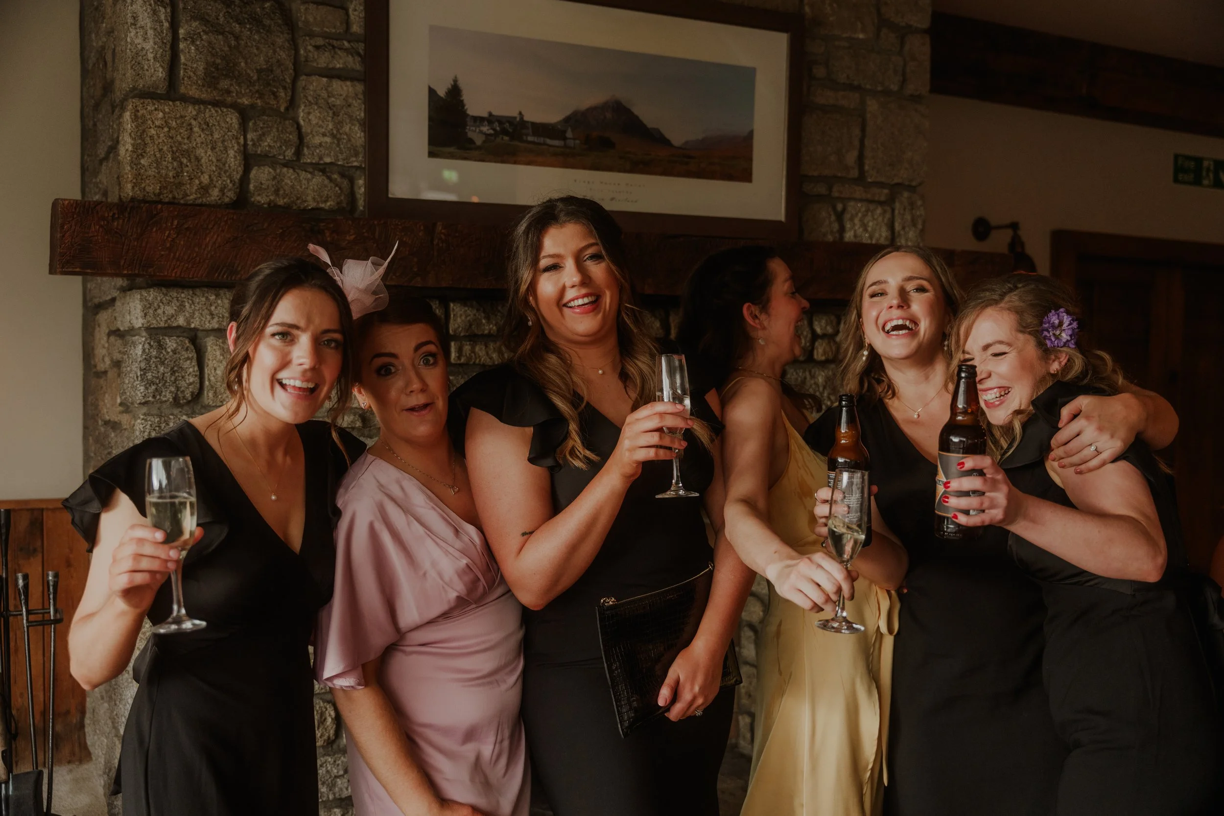 Six women in elegant dresses celebrating together indoors, holding drinks, smiling and laughing.
