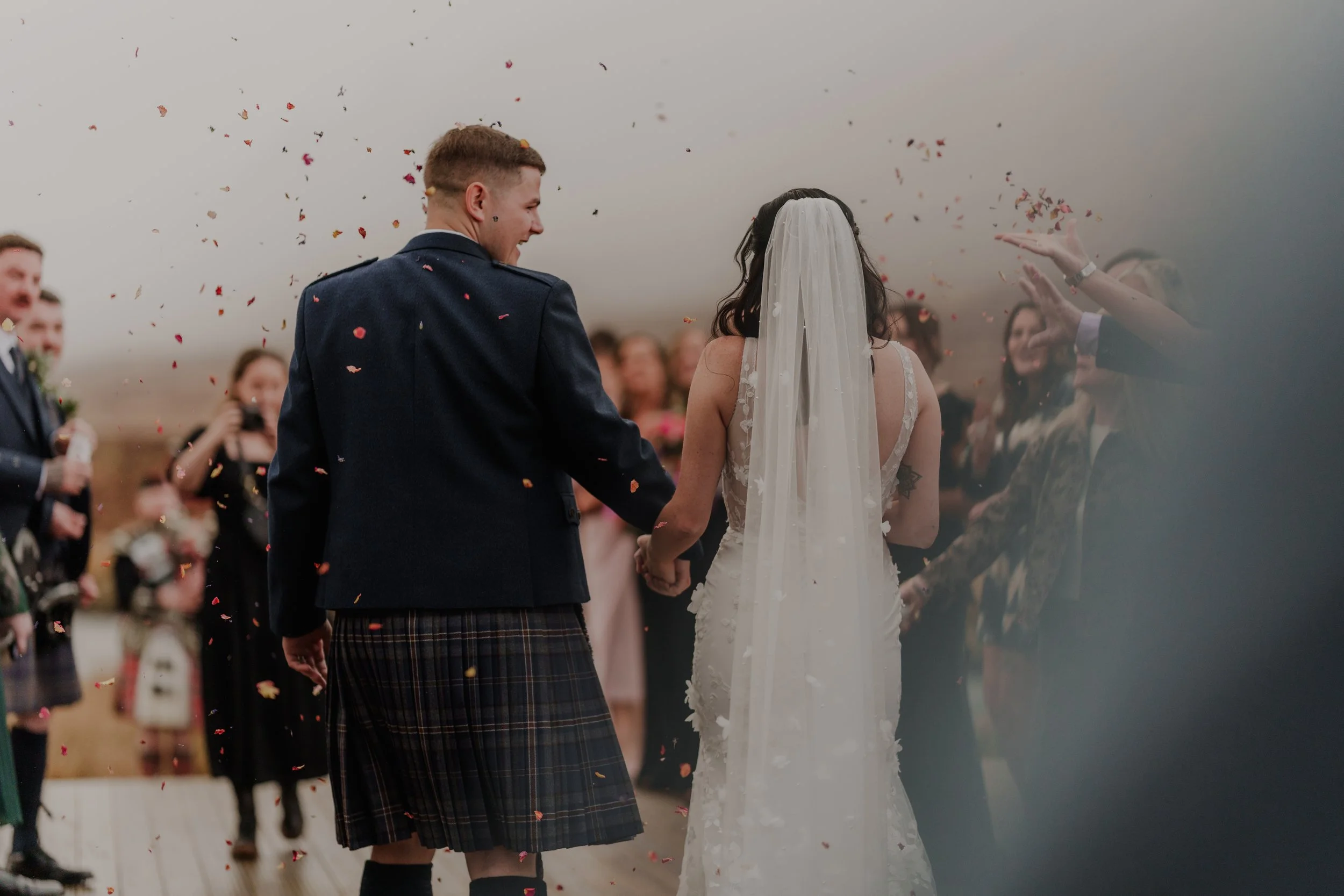 A bride and groom holding hands at their wedding reception as people toss confetti in the air.