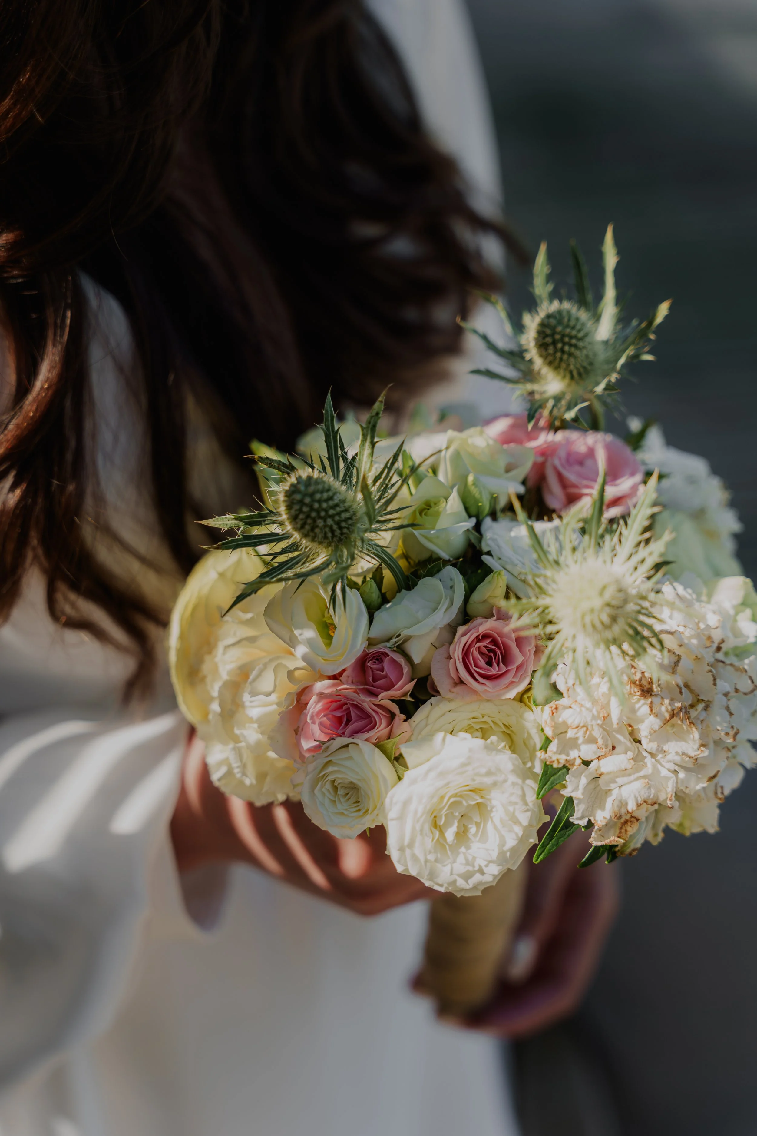 Close-up of a woman holding a bouquet of pink, white, and cream roses, along with other flowers and greenery, with a blurred dark background.