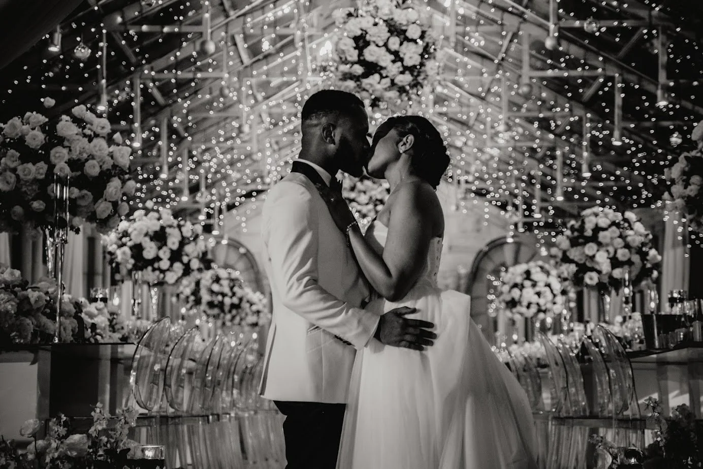 A black and white photo of a bride and groom kissing at their wedding reception, decorated with floral arrangements and string lights overhead.