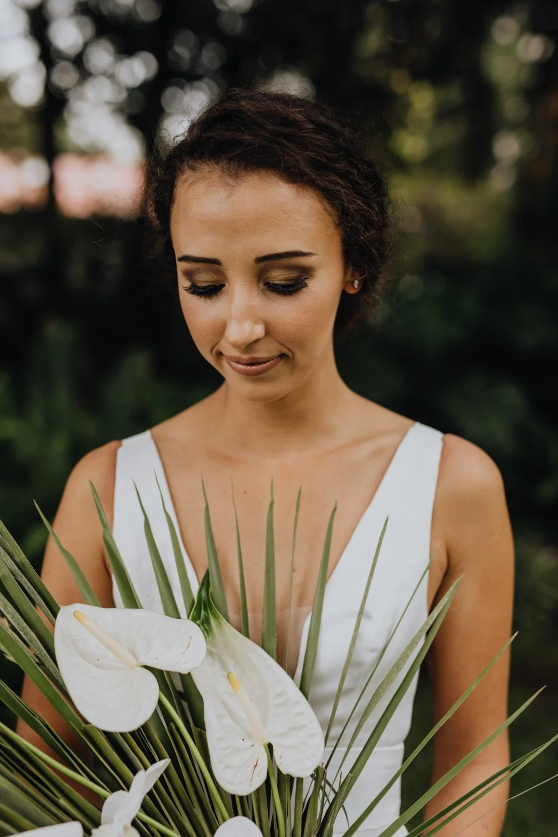 A woman in a white dress holding a bouquet of white calla lilies and green foliage outdoors.