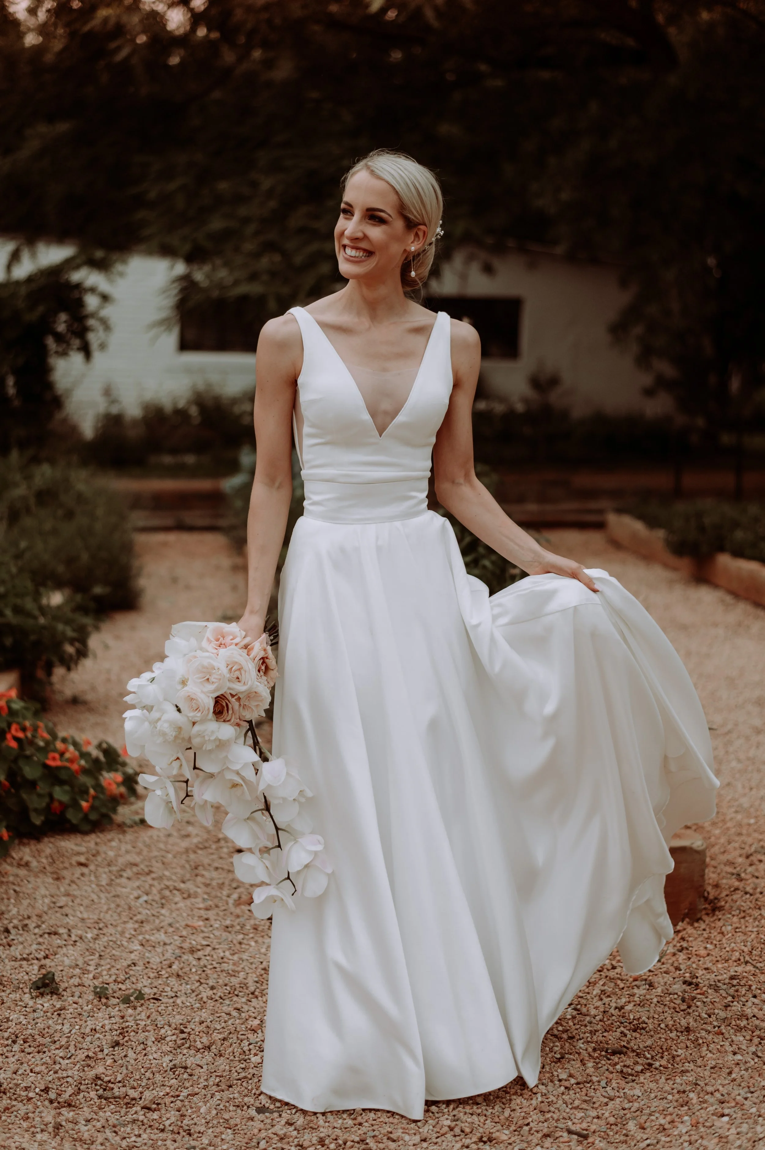 A smiling bride in a white wedding dress holding a bouquet of pink and white roses and orchids, walking outdoors on a gravel pathway with greenery around.