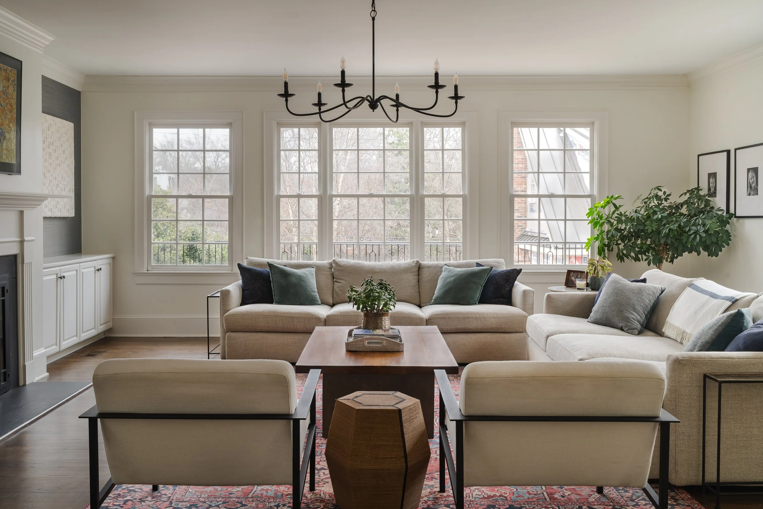 A living room with beige sofas, green and gray pillows, a wooden coffee table, and a large window with a view of trees outside. There is a tall houseplant on the right, framed pictures on the wall, and a black chandelier hanging from the ceiling.