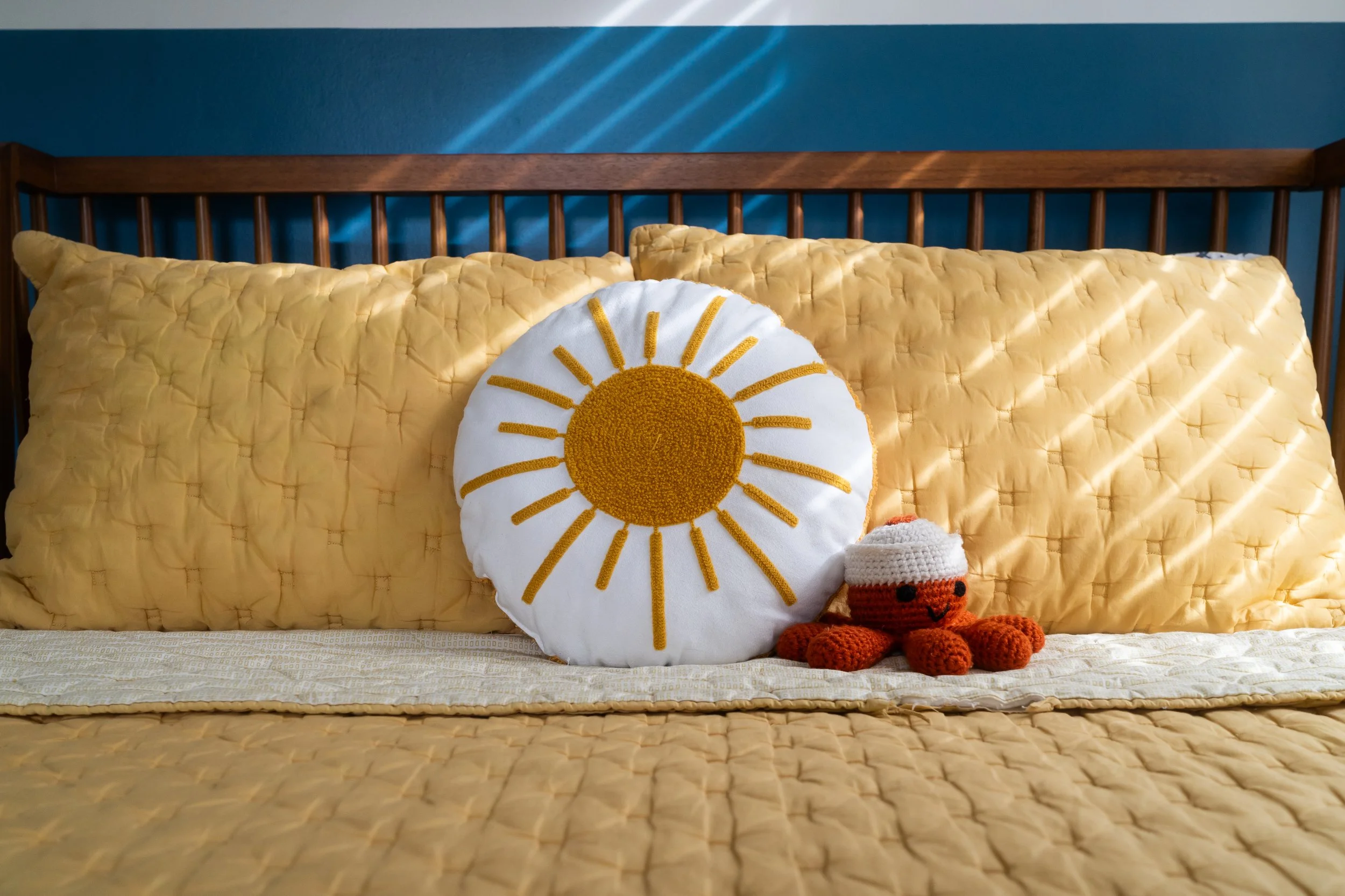 A bed with yellow quilted pillows, a decorative pillow with a sun design, and a small crocheted orange teddy bear, set against a teal wall with sunlight streaming through blinds.