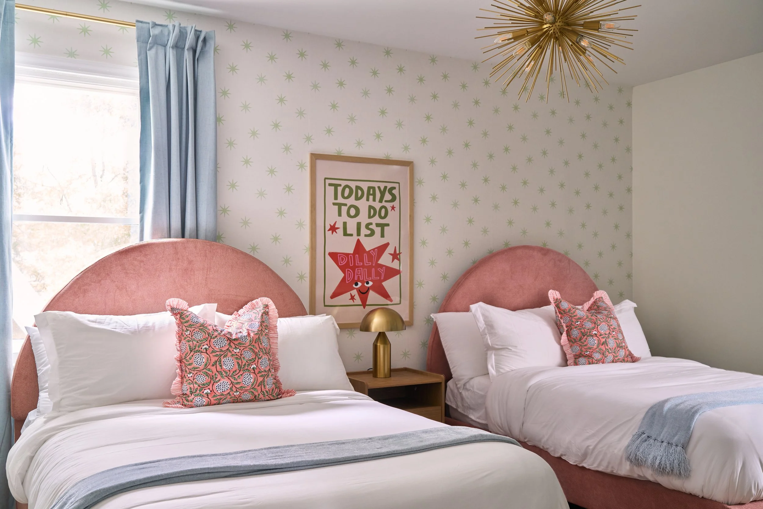two beds with pink velvet headboards in a playful bedroom with green star wallpaper and blue curtains