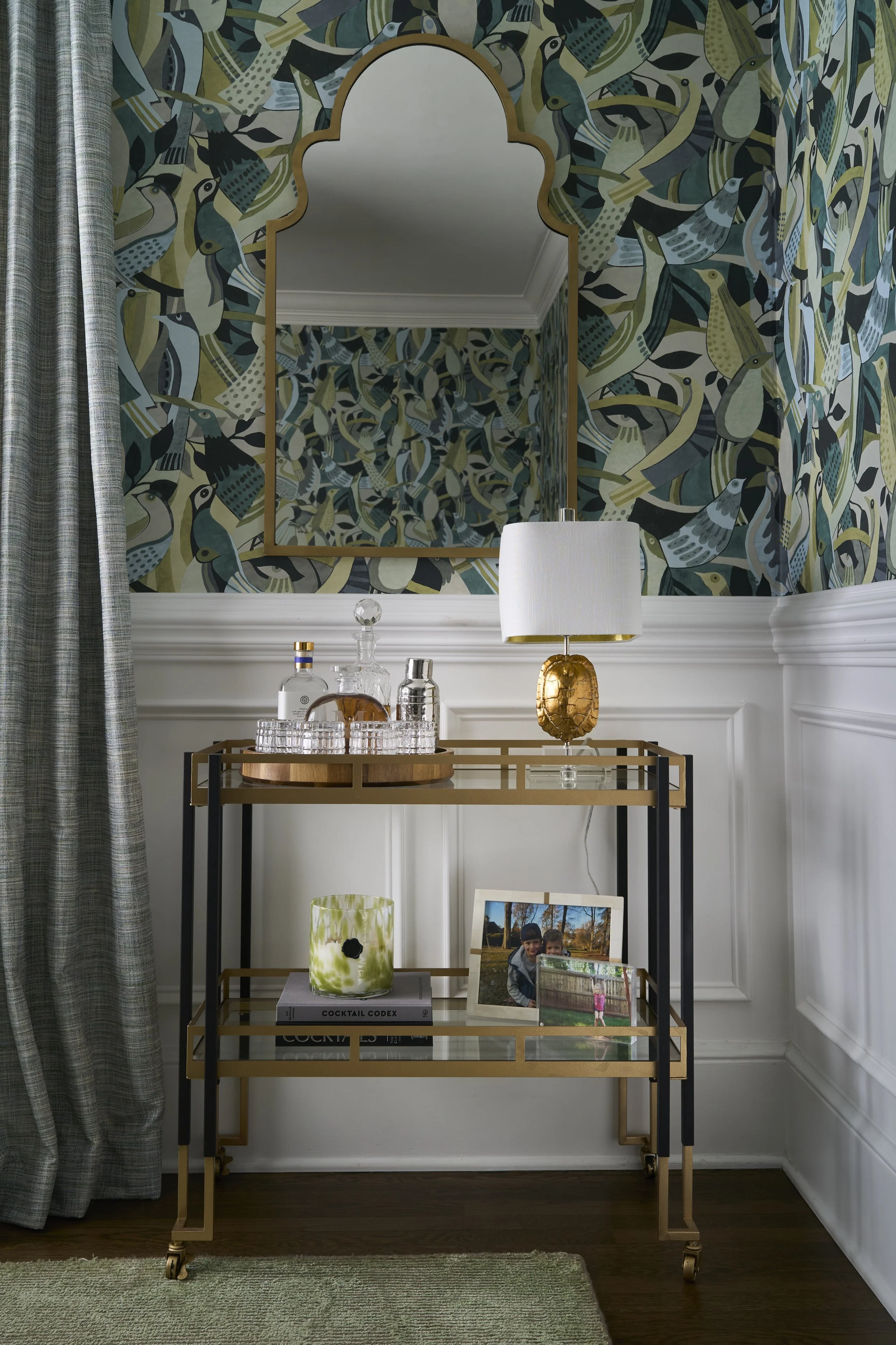 A gold and black metal bar cart holding glass decanters, a candle, books, and framed photographs, situated in a room with green and blue bird-patterned wallpaper and a large mirror.