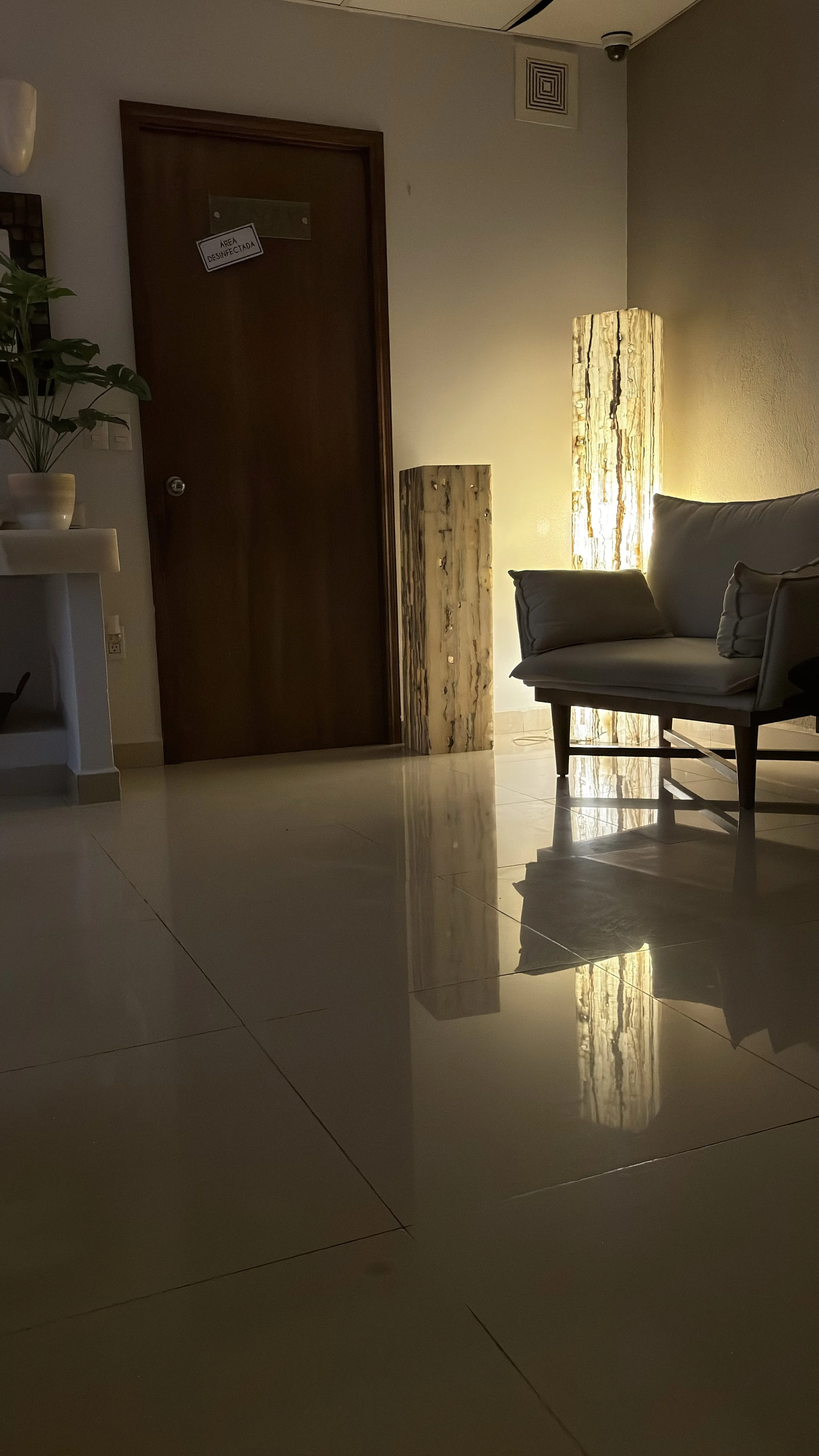 Sala de espera con lámpara decorativa y un sillón, puerta con señal de área desinfectada, planta en maceta, suelos de baldosas blancas y paredes beige.