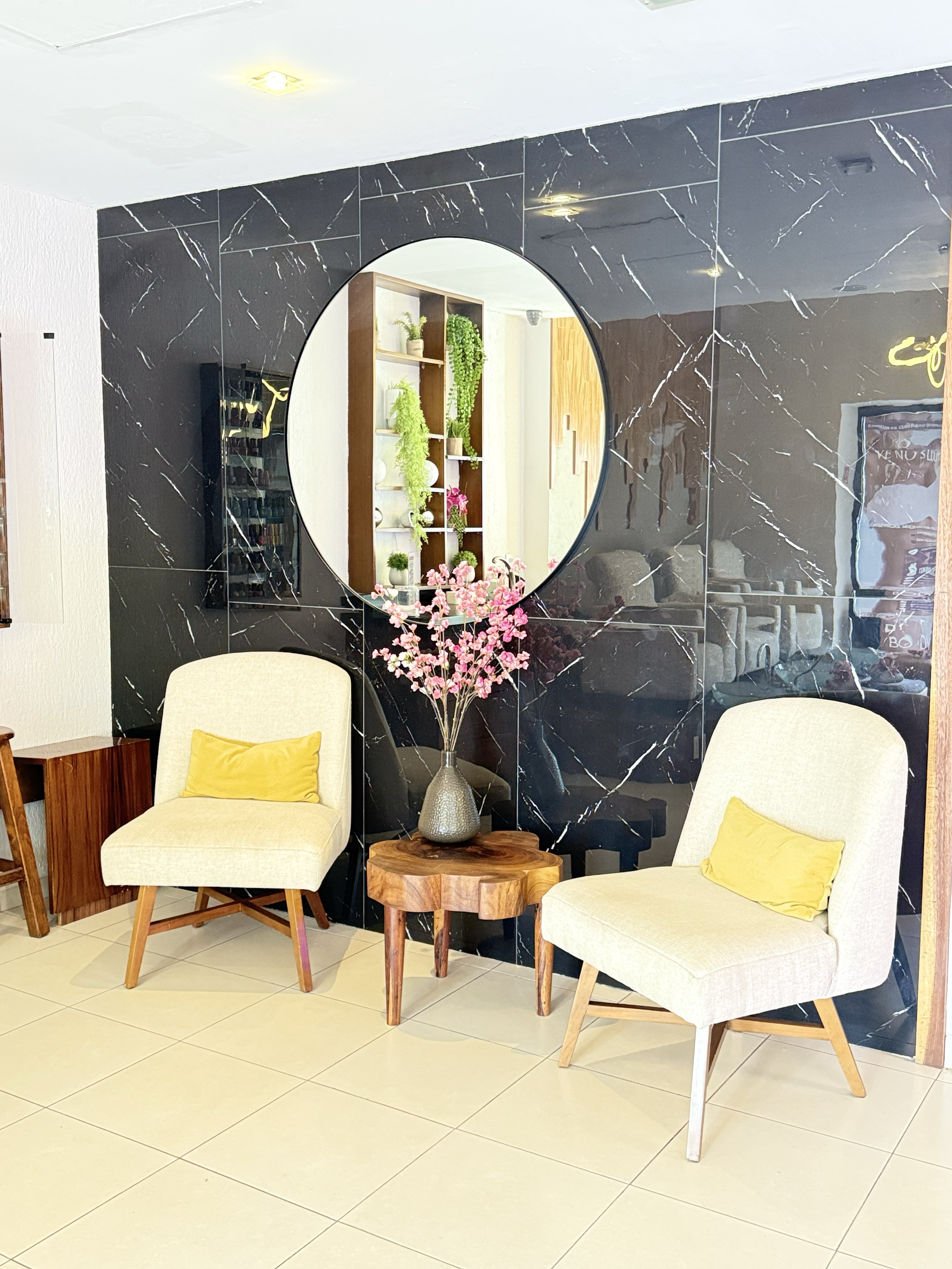 Sala de estar con sillones beige, cojines amarillos, mesa de madera, florero con flores rosas, pared decorativa negra con mármol, espejo redondo, estantería con plantas y objetos decorativos, paredes blancas y piso de baldosas blancas.