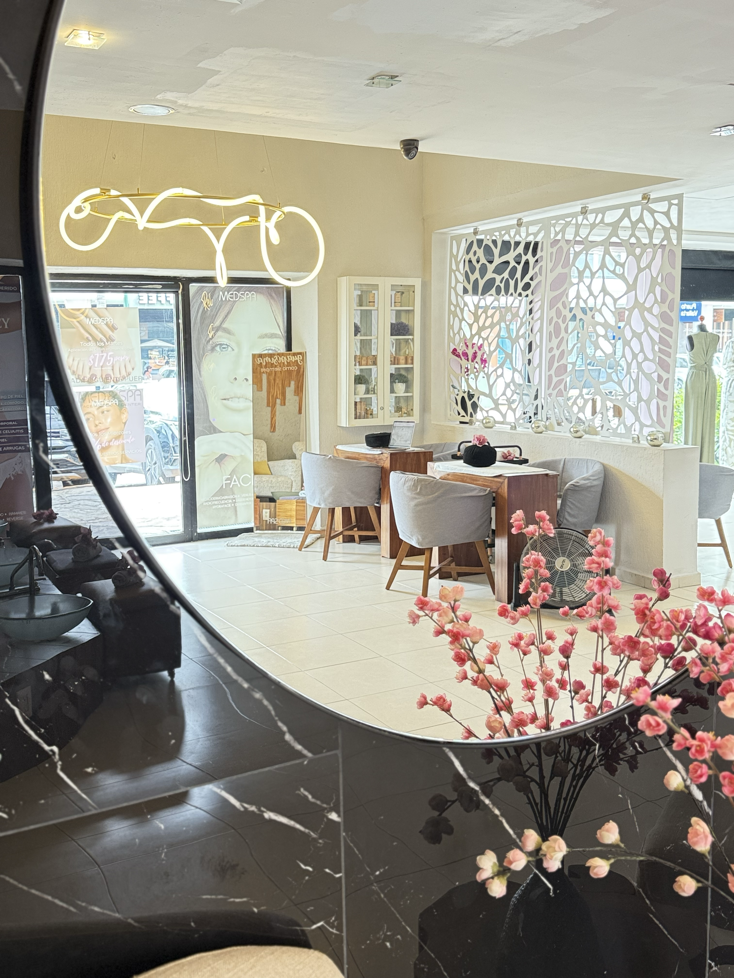 Vista del interior de un salón con mesas y sillas donde hay una decoración moderna y flores artificiales en una mesa. Se puede ver una pantalla de promoción en la entrada, una iluminación decorativa en forma de bicicleta y una estructura de celosía blanca que divide el espacio.