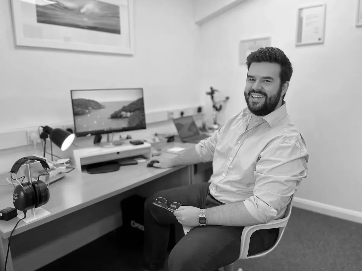 A man sitting at a desk in an office with a computer monitor, headphones, and various office supplies, smiling and holding glasses.