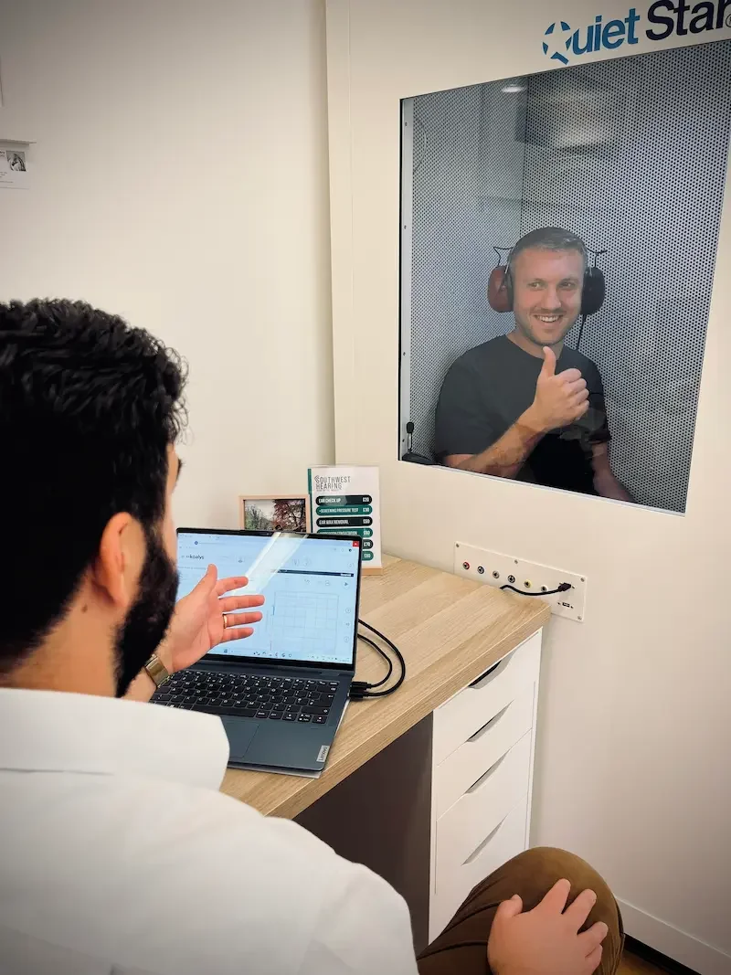 Man with headphones giving a thumbs up inside an audiology booth while an audiologist interacts with a computer in a hearing test appointment.