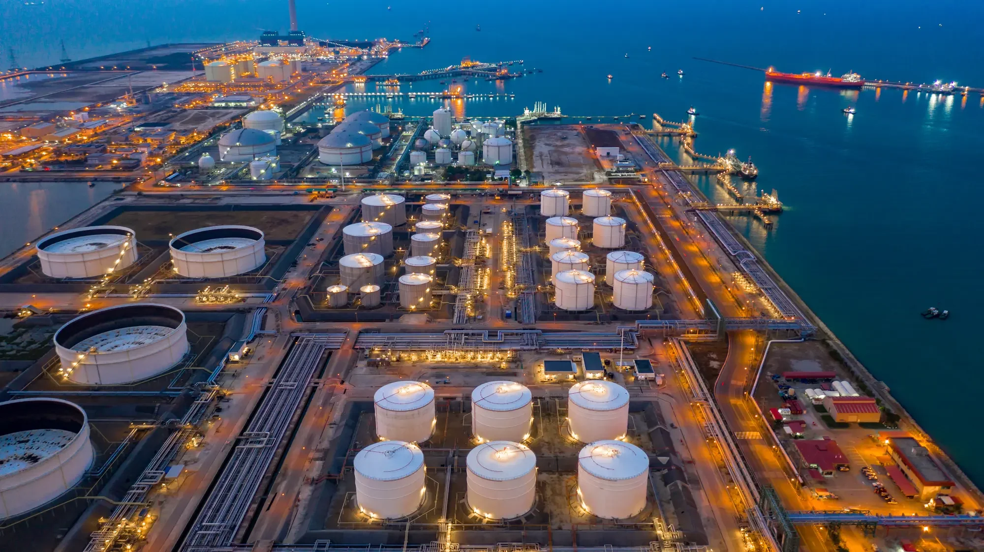 Oil refinery at night featuring storage tanks and water, highlighting the infrastructure of oil and gas facilities