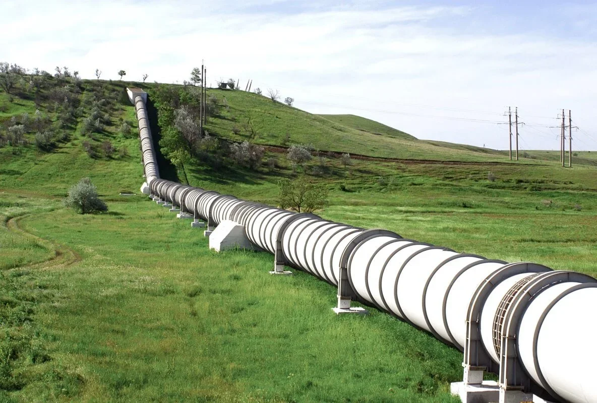 A large gas pipeline stretches across a vibrant green field under a clear blue sky