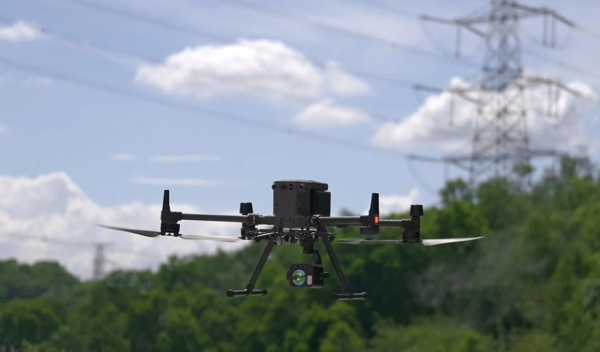 A drone with a methane detection payload hovers above a field, surrounded by power lines under a clear sky