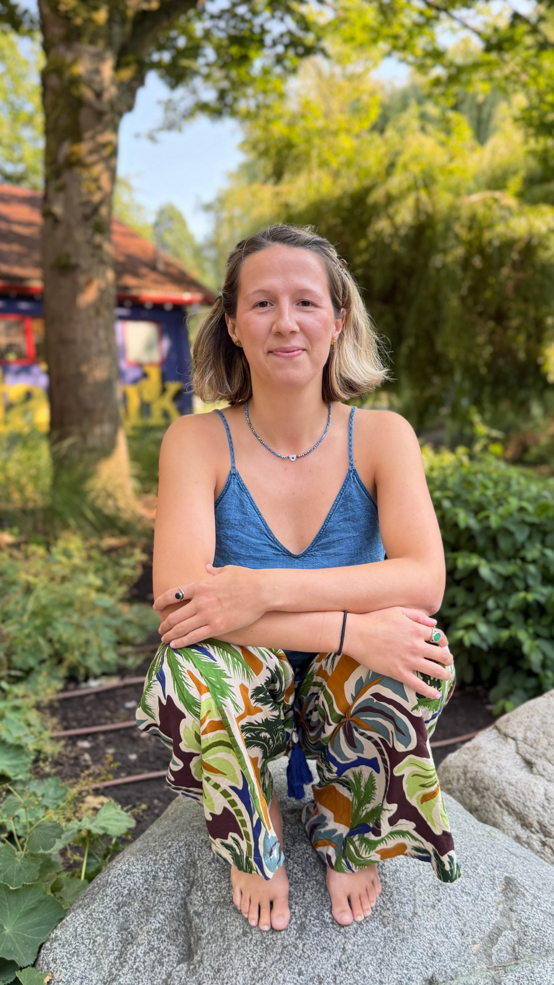 Dr Alex Dragan in a blue spaghetti strap top and colorful patterned pants is sitting barefoot on a rock in a garden with greenery and trees in the background.
