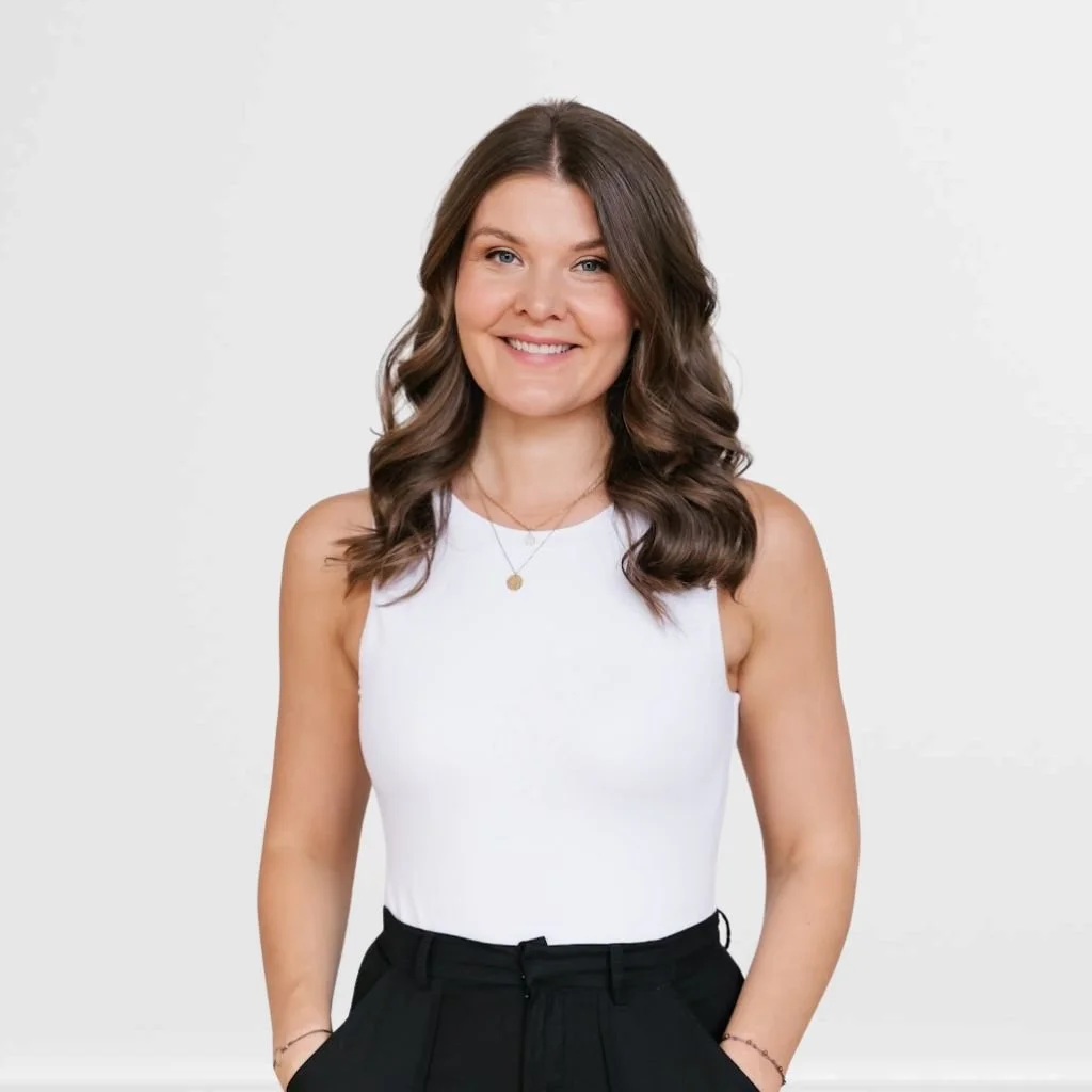 A woman with wavy brown hair wearing a white sleeveless top and black pants, smiling against a plain white background.