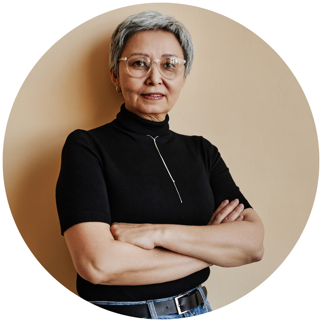 An older woman with short gray hair wearing glasses and a black turtleneck, standing with her arms crossed against a beige wall.