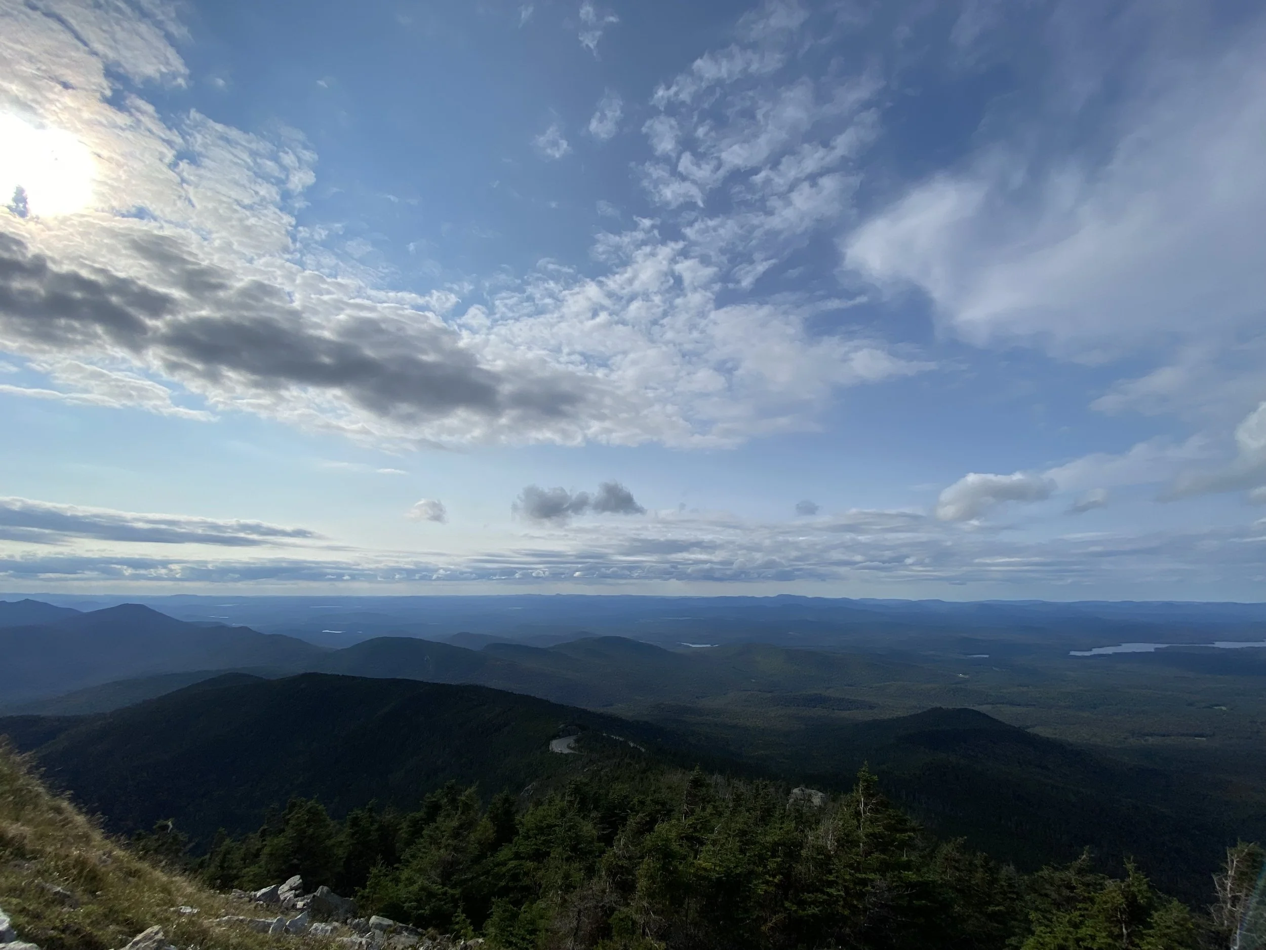 Scenic mountain landscape with cloudy sky and sun peeking through, featuring lush green forests and rolling hills in the distance.