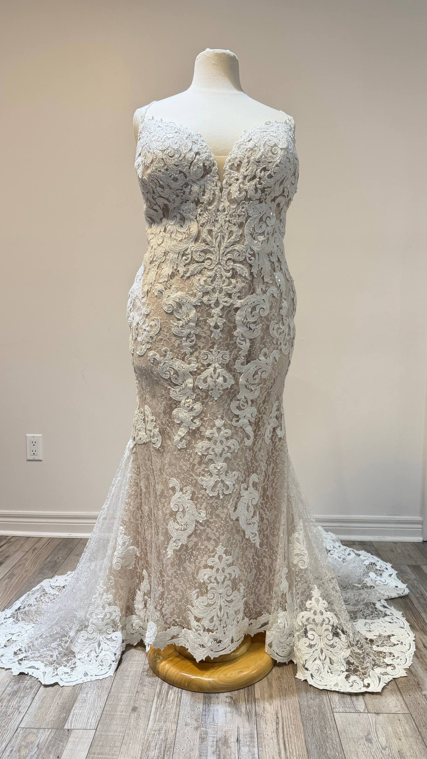 A mannequin displays an elegant, sleeveless, ivory lace wedding gown with intricate floral embroidery, fitted silhouette, and a train on a wooden floor against a plain wall.