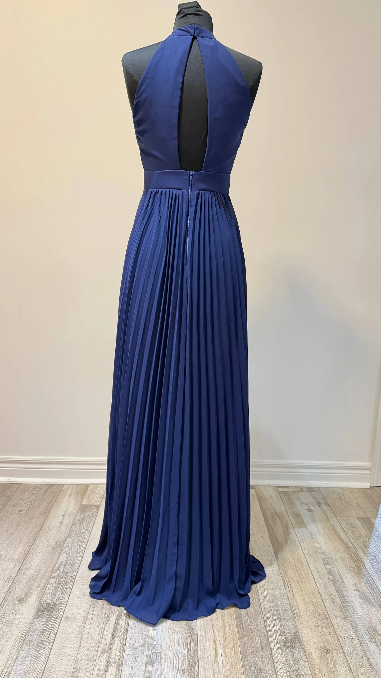 A royal blue evening gown with a pleated floor-length skirt and a halter neckline, displayed on a black mannequin against a plain beige wall.