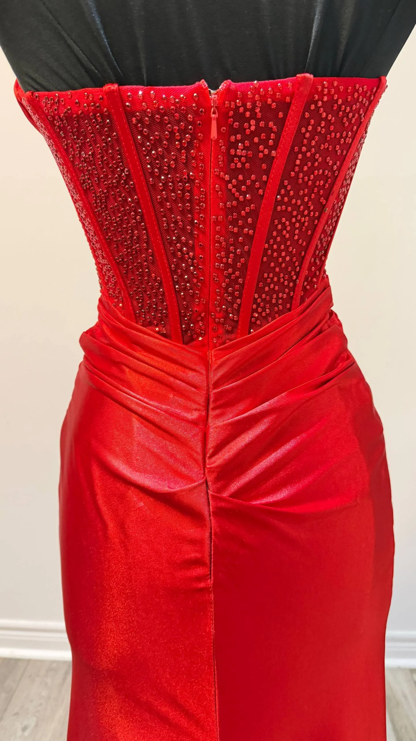 A woman wearing a fitted red dress with a high waist and a detailed red corset with embellishments, with a black top or jacket visible at the top of the image.