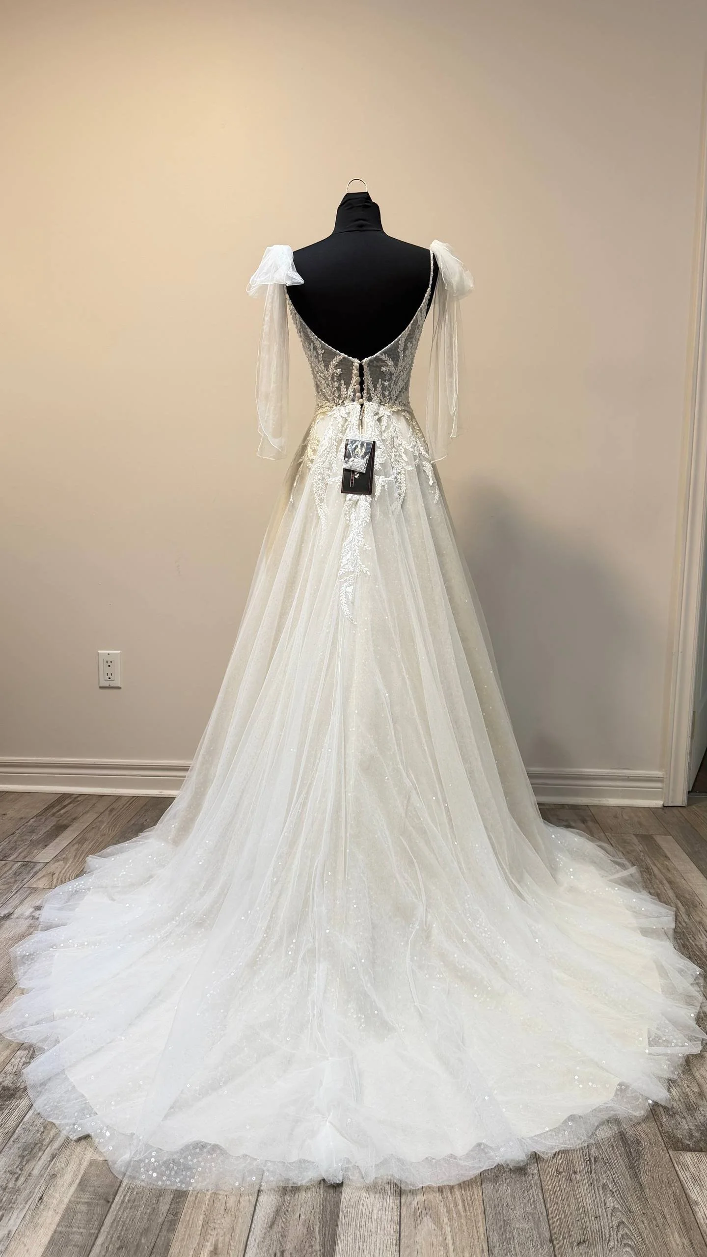 A wedding dress on a mannequin, featuring a fitted bodice with lace details and a full, flowing tulle skirt with subtle sparkle, displayed indoors against a plain wall.