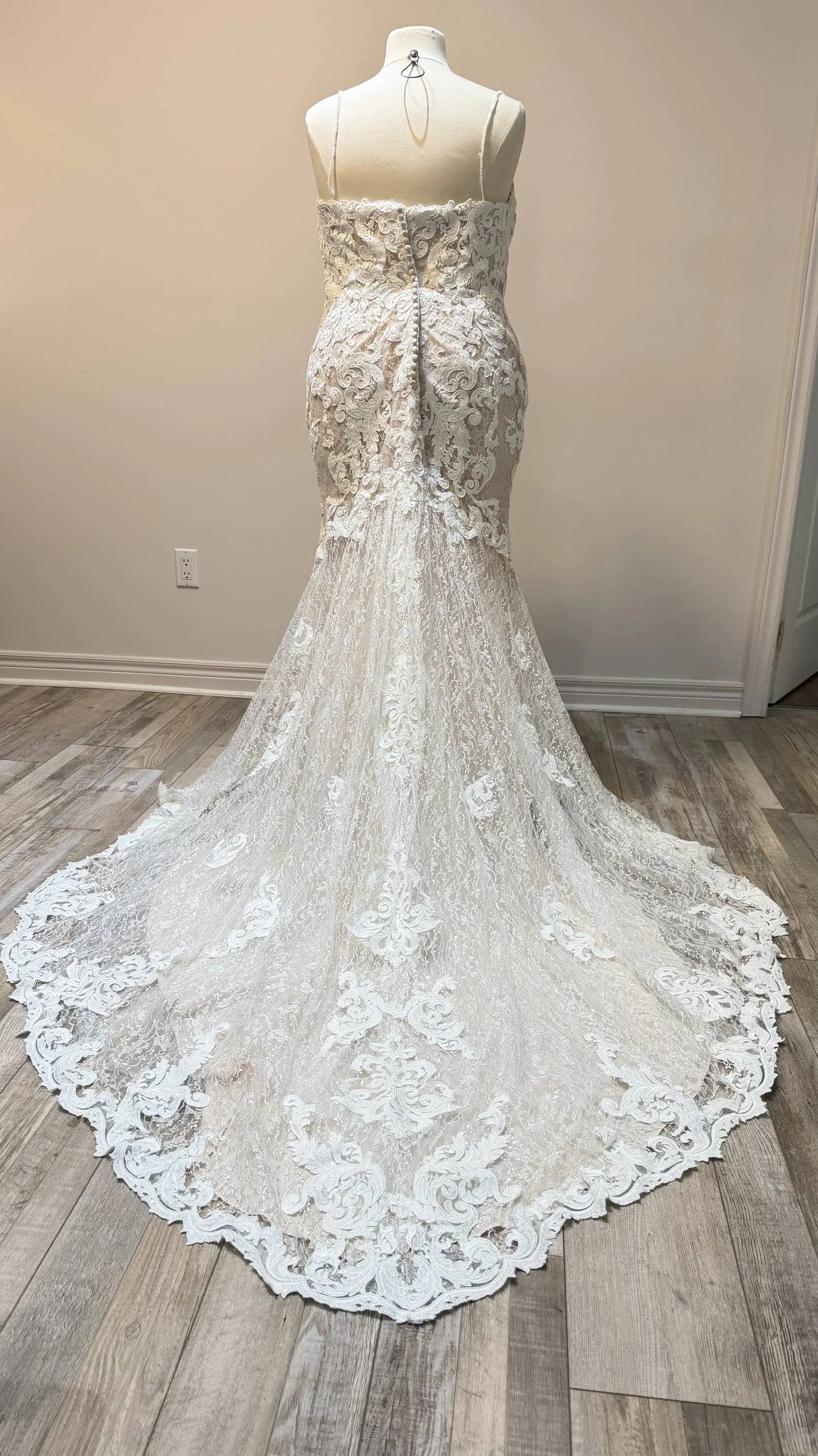 White lace wedding dress on a mannequin with a long train and spaghetti straps displayed indoors.