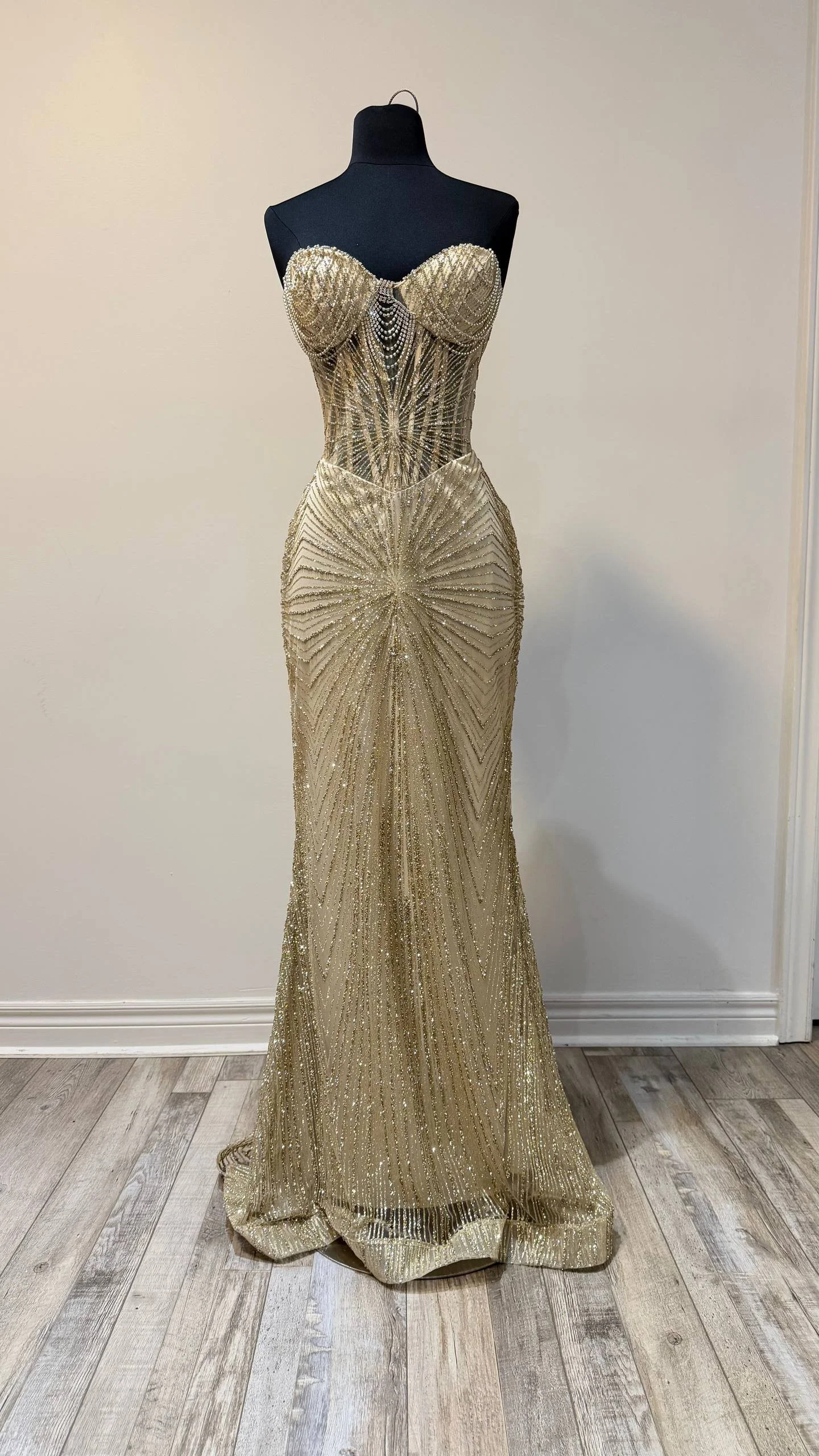A luxurious gold evening gown with intricate beading and embellishments displayed on a black mannequin against a plain wall.