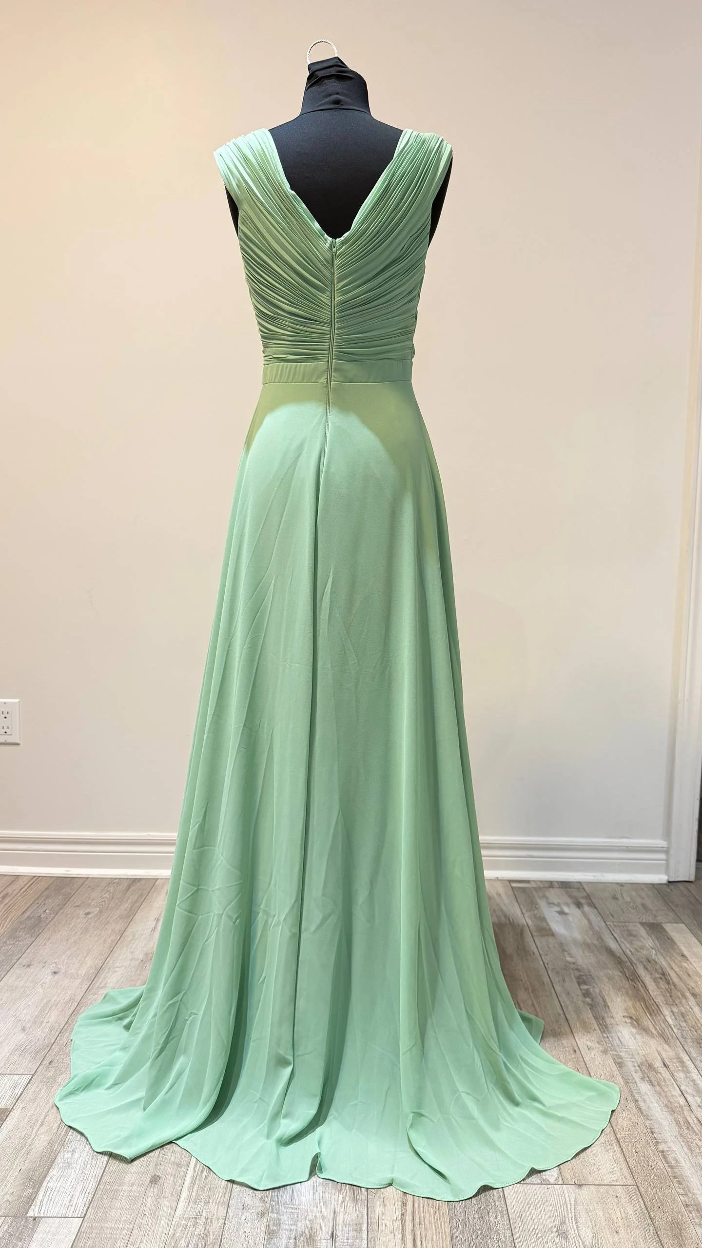 A long, flowing, light green gown with a ruched bodice and deep V-neckline, displayed on a black dress form against a plain wall.