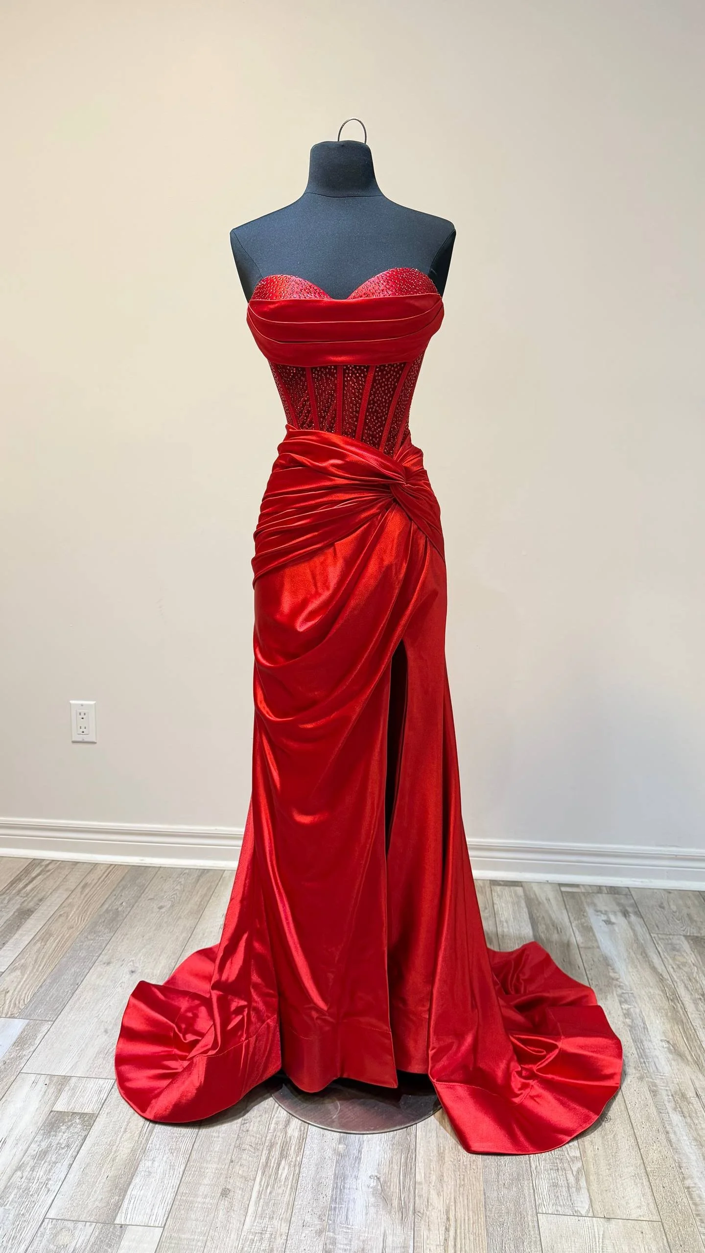 Red evening gown displayed on a black mannequin, featuring a strapless, fitted bodice with embellishments, and a long flowing skirt with a front slit, gathered and draped at the waist.